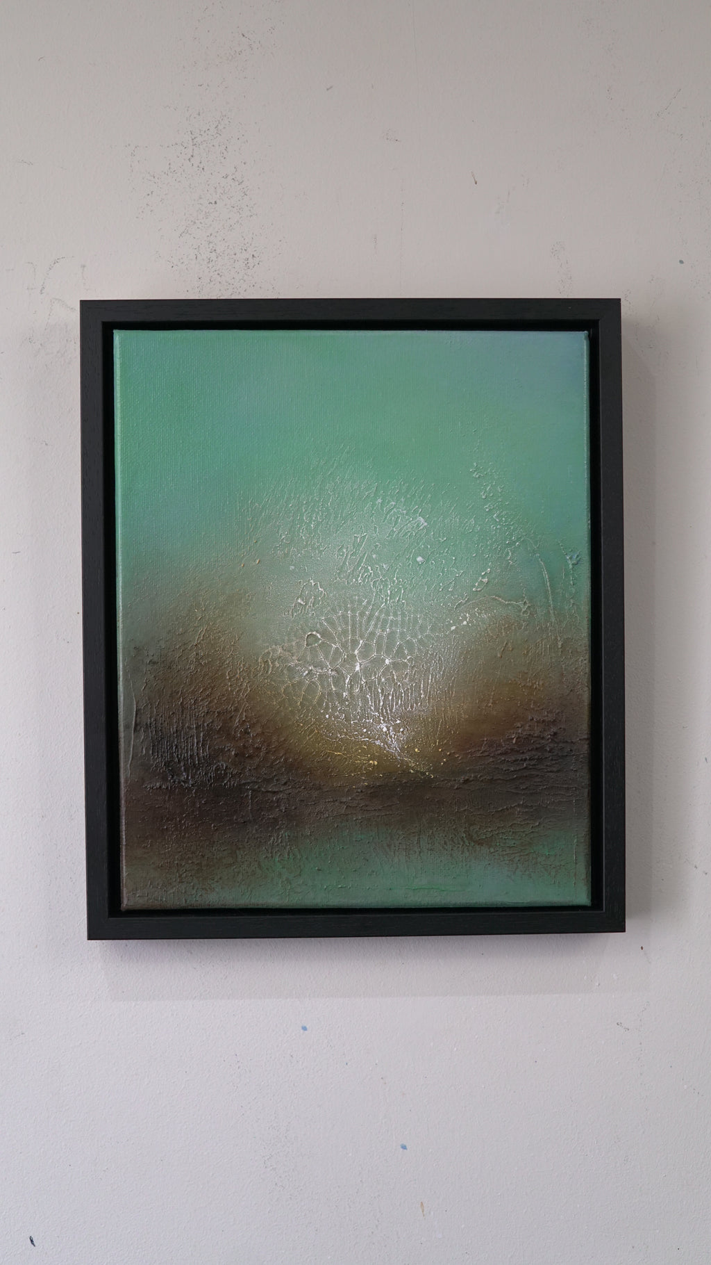 A framed abstract painting featuring a textured teal and brown landscape with a bright white glowing center, housed in a black frame and mounted on a white wall.