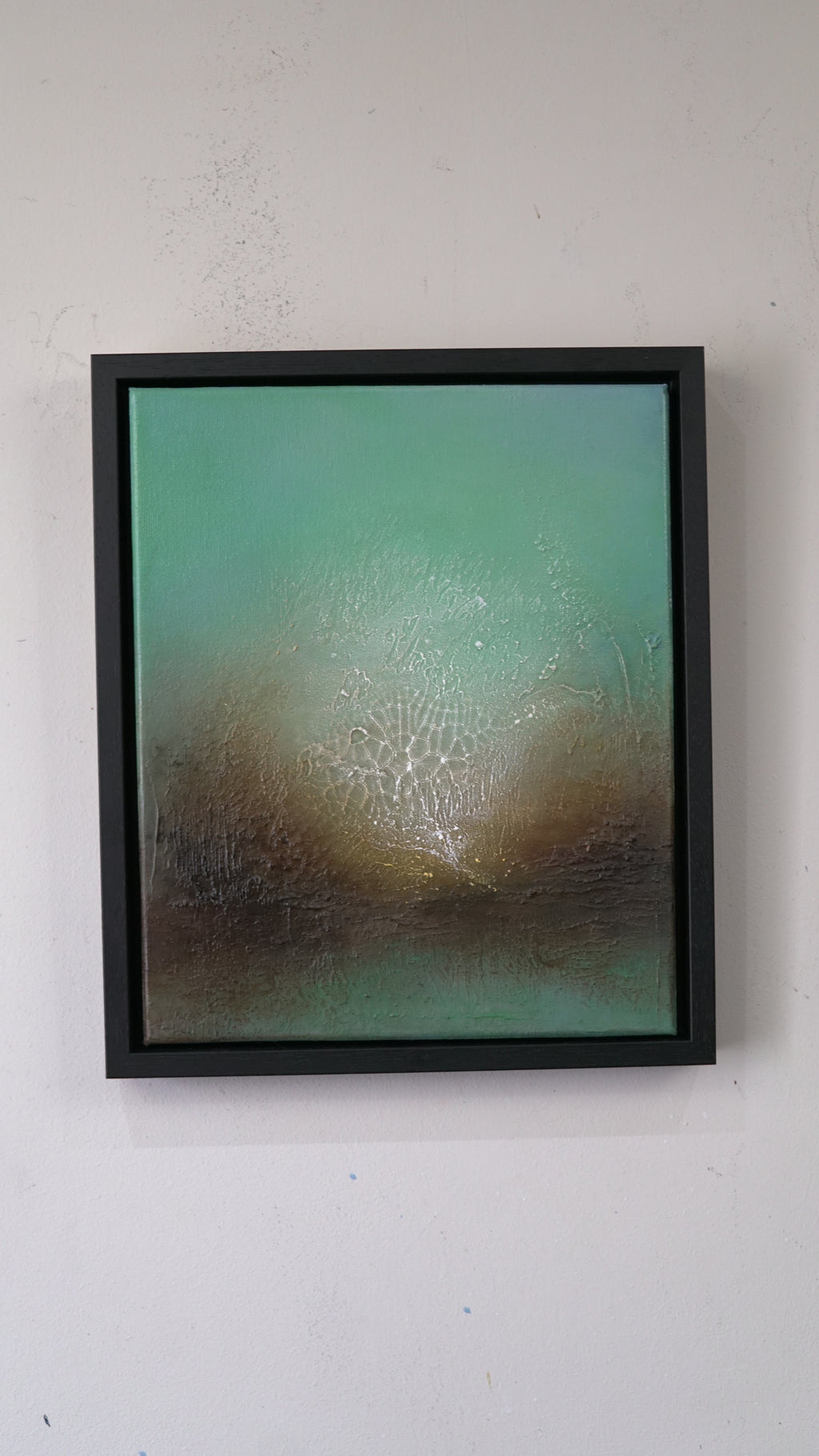 A framed abstract painting featuring a textured teal and brown landscape with a bright light source, mounted on a plain white wall.