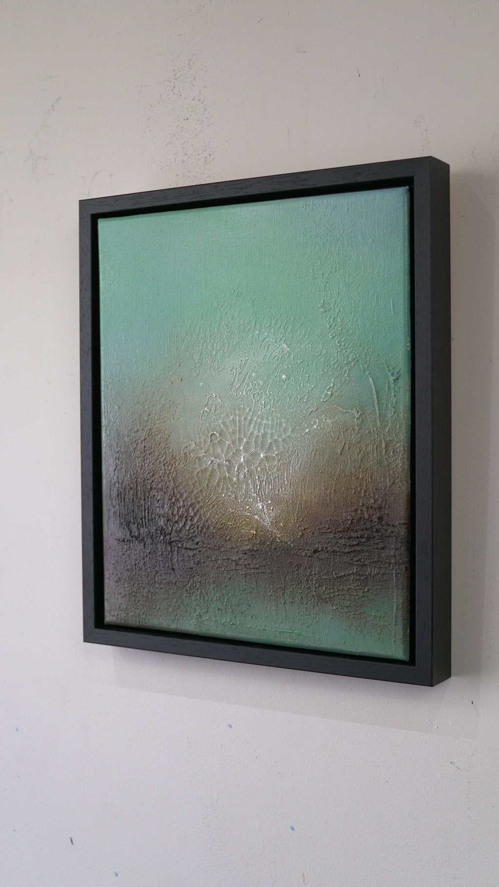 A framed abstract wall art piece hanging on a white wall. The artwork features a textured gradient transitioning from a soft mint green at the top to earthy brown and gray tones near the bottom, enclosed in a thick black frame.