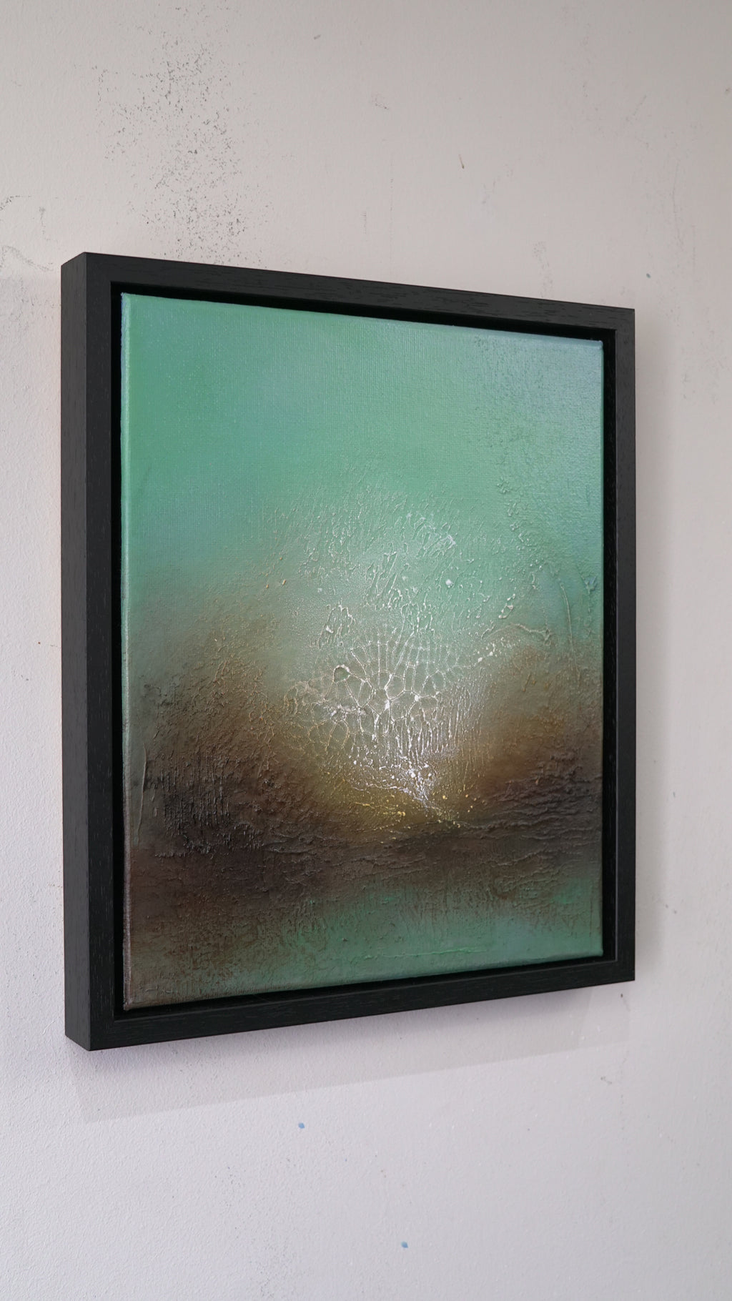 A framed abstract canvas painting hanging on a white wall. The artwork features a textured gradient of teal green at the top blending into earthy brown tones at the bottom, enclosed in a thick black frame.
