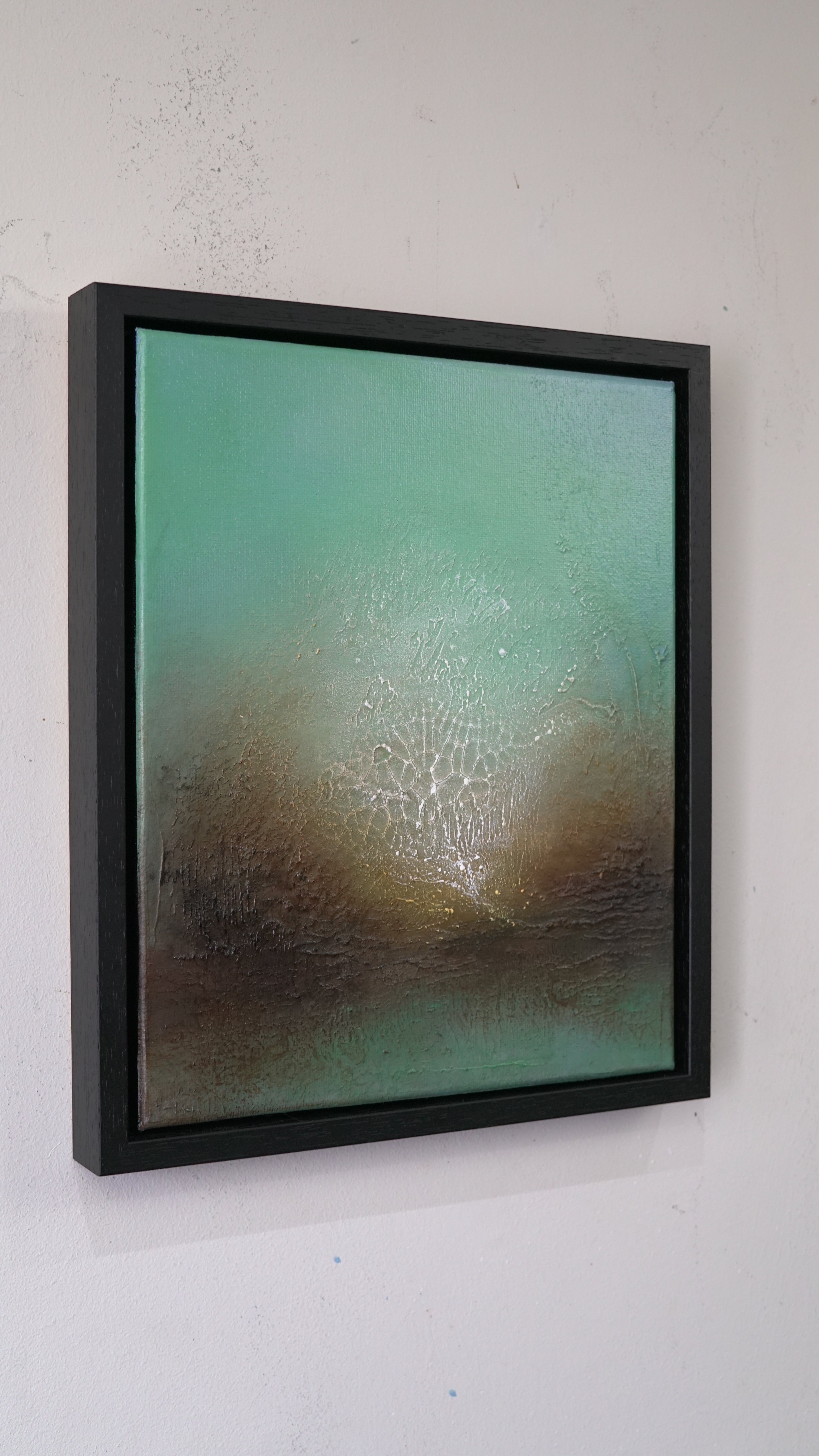 A framed abstract canvas painting hanging on a white wall. The artwork features a textured gradient of teal green at the top blending into earthy brown tones at the bottom, enclosed in a thick black frame.