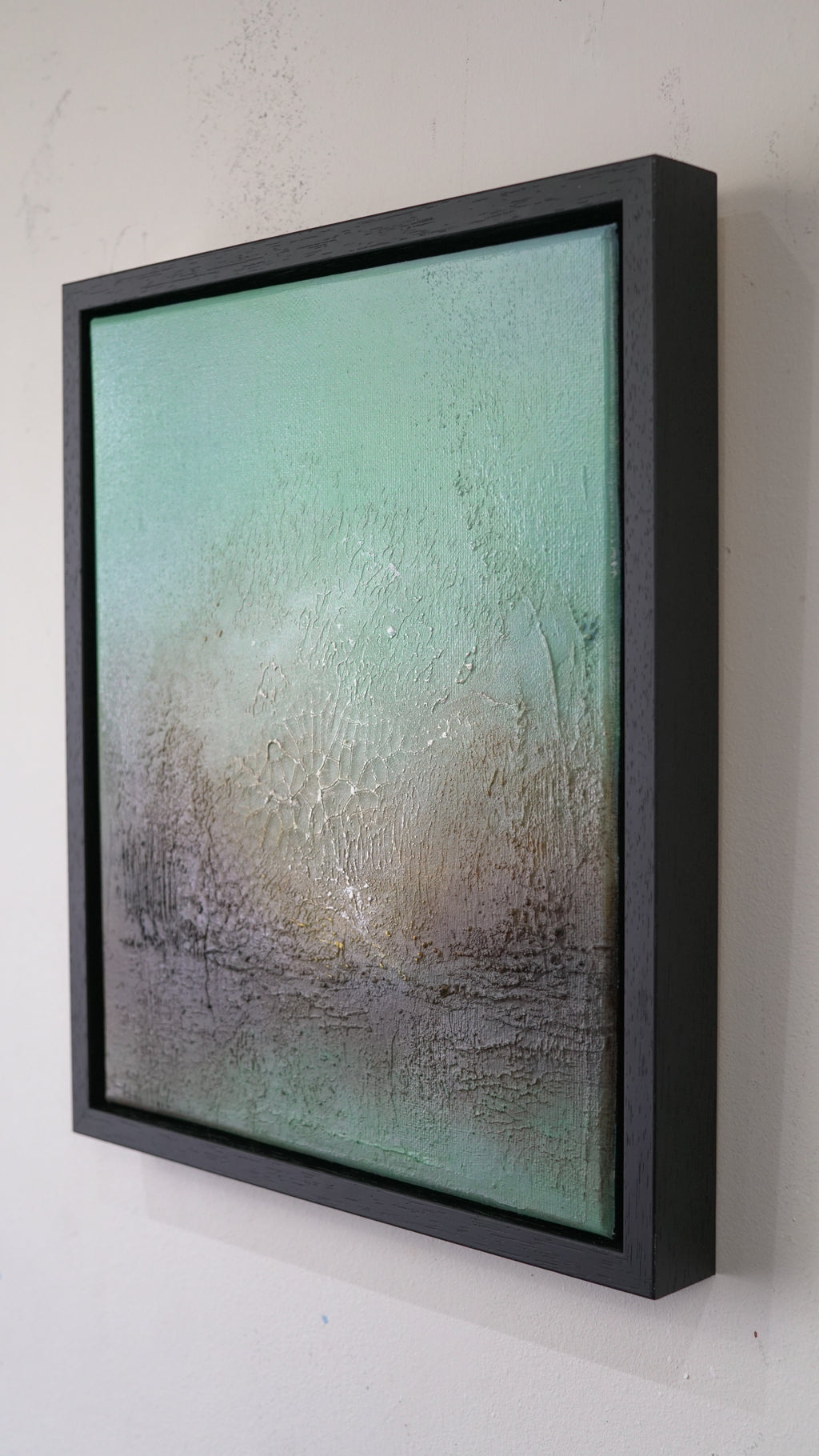 A framed abstract canvas print featuring a textured gradient of mint green, beige, and gray, housed in a thick black float frame and mounted on a white wall.