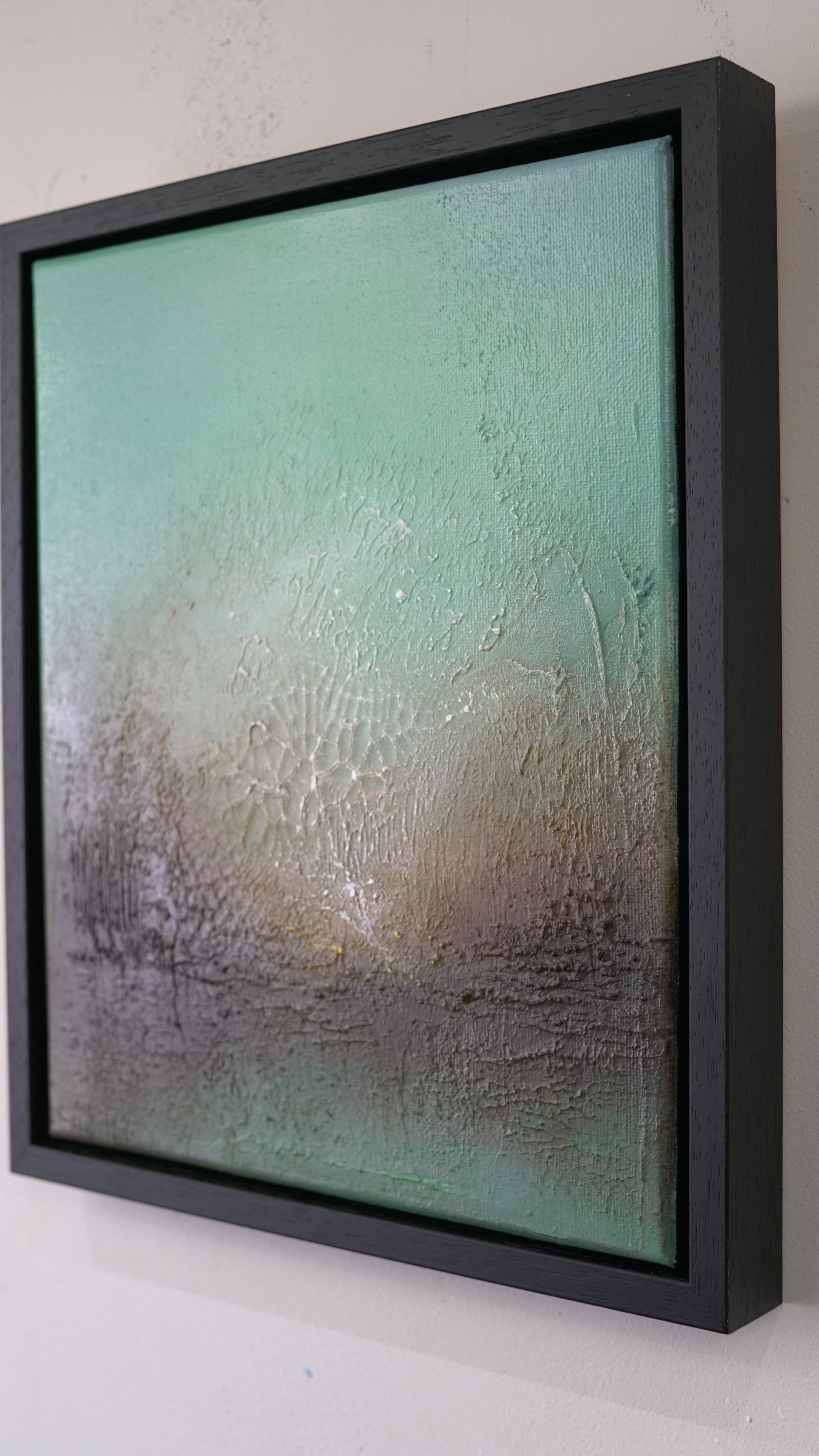 A square framed abstract painting with a textured finish, featuring a gradient of mint green, beige, and gray tones, mounted on a white wall.
