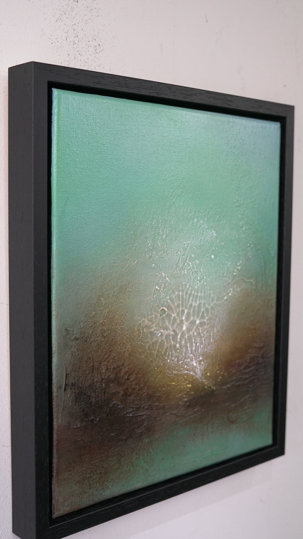 Framed abstract wall art hanging on a white wall, featuring a textured teal and brown composition with a black frame.