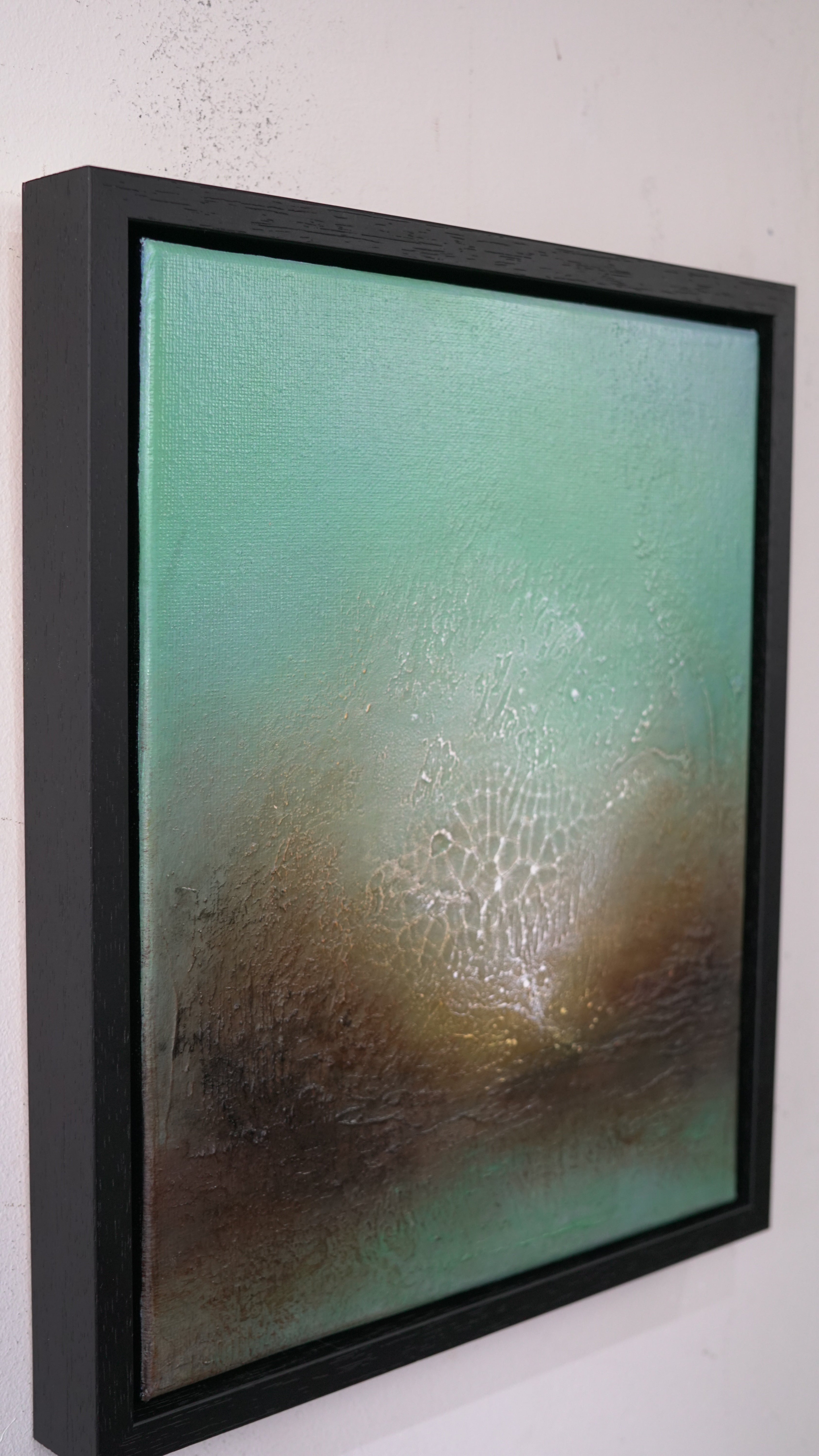 Framed abstract wall art hanging on a white wall, featuring a textured teal and brown composition with a black frame.