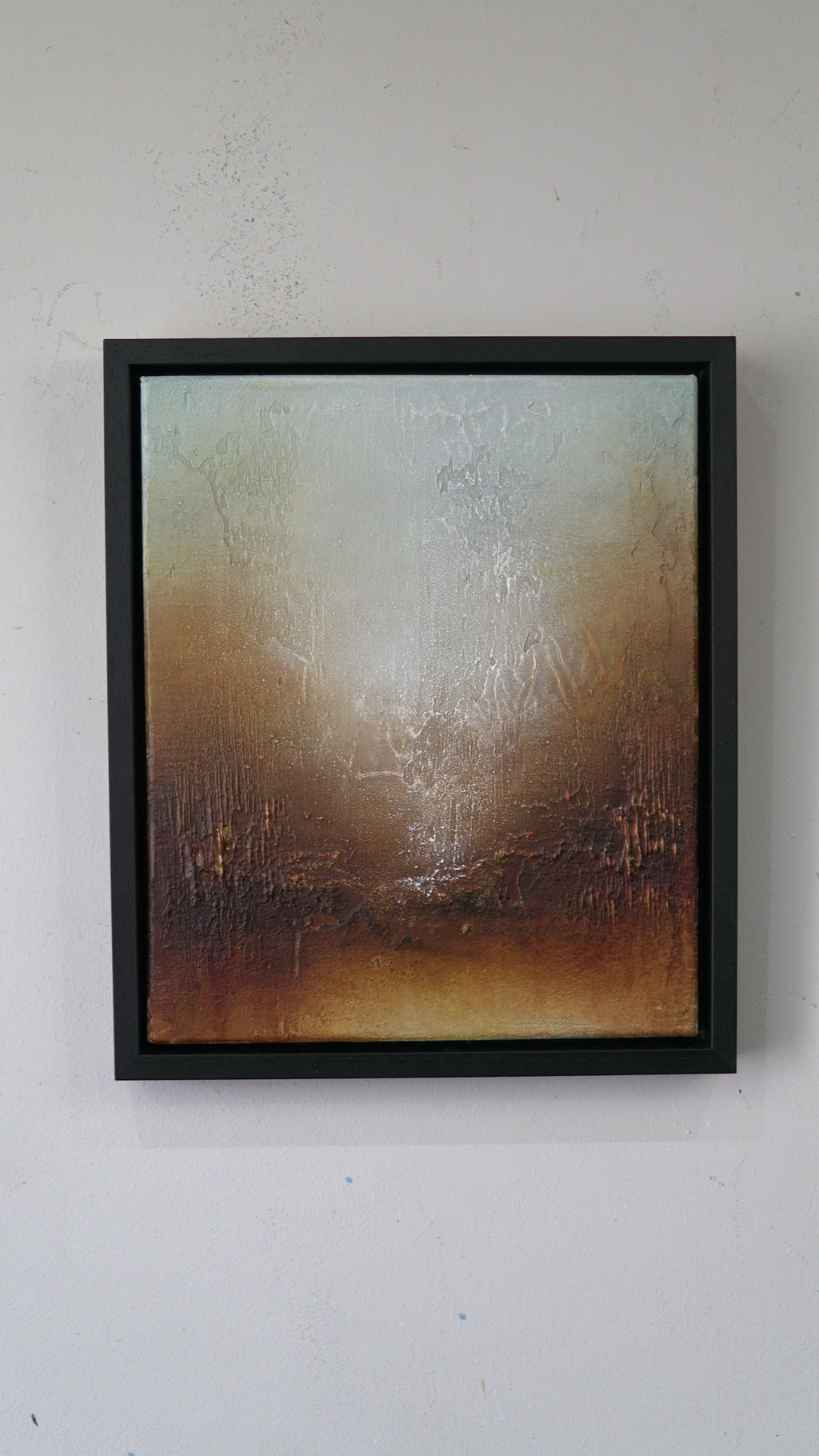 A framed abstract painting with a textured, distressed finish in earthy tones of brown, beige, and muted blue-gray, housed in a simple black frame and mounted on a white wall.