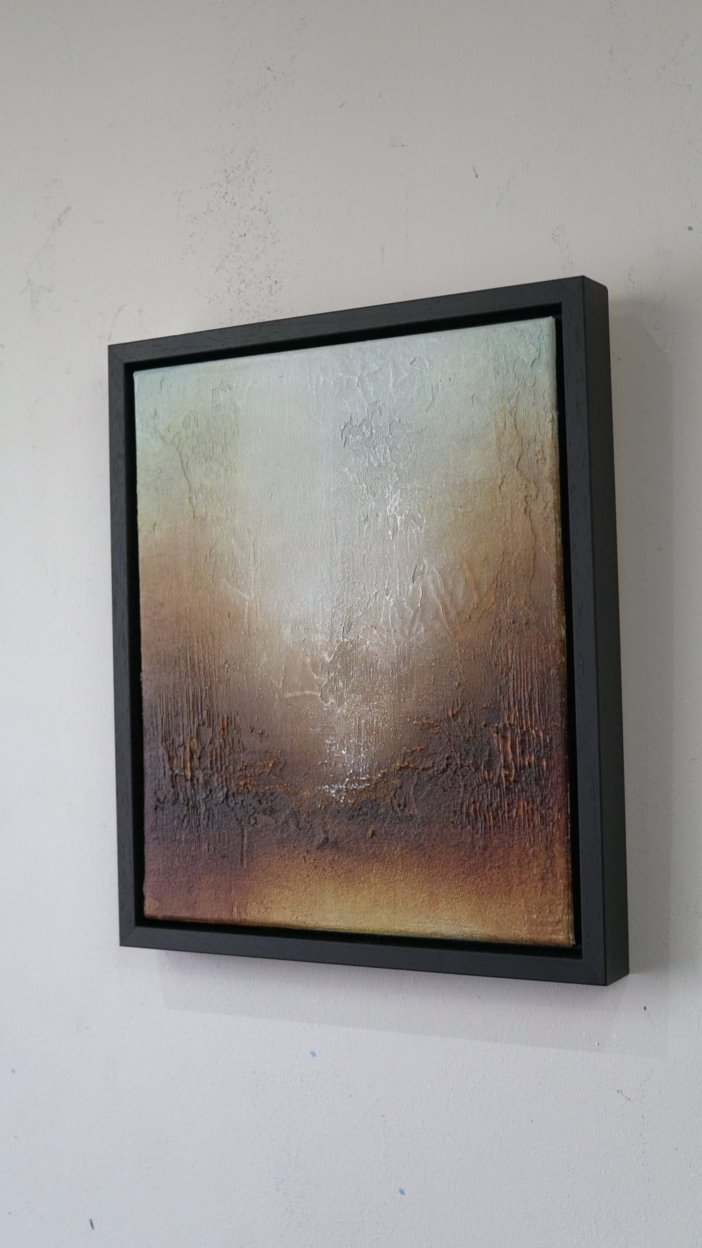 A framed abstract canvas painting with a textured, distressed finish in earthy tones of brown, beige, and gray, housed in a thick black frame and mounted on a white wall.