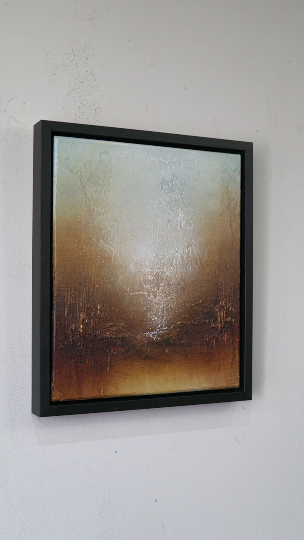A framed abstract painting with textured brushstrokes in warm brown, beige, and muted blue-gray tones, displayed on a white wall.