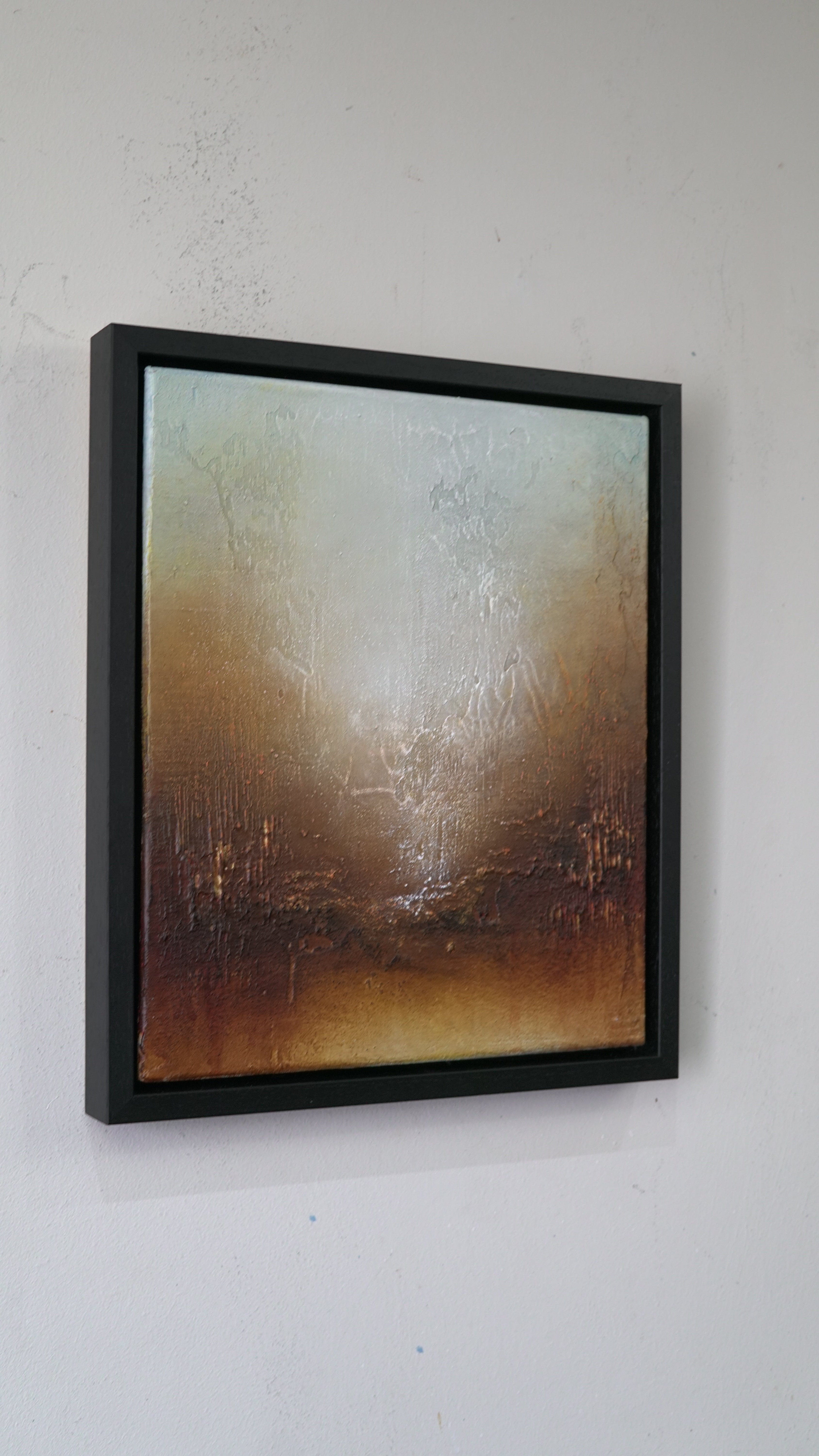 A framed abstract painting with textured brushstrokes in warm brown, beige, and muted blue-gray tones, displayed on a white wall.