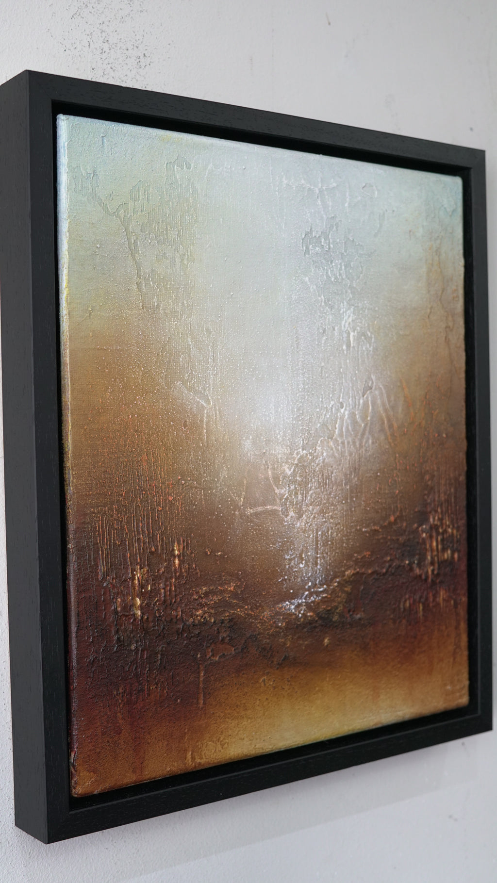 Framed abstract canvas wall art hanging on a white wall, featuring a textured gradient from pale cream and silver at the top to warm brown and bronze tones at the bottom, enclosed in a black frame.