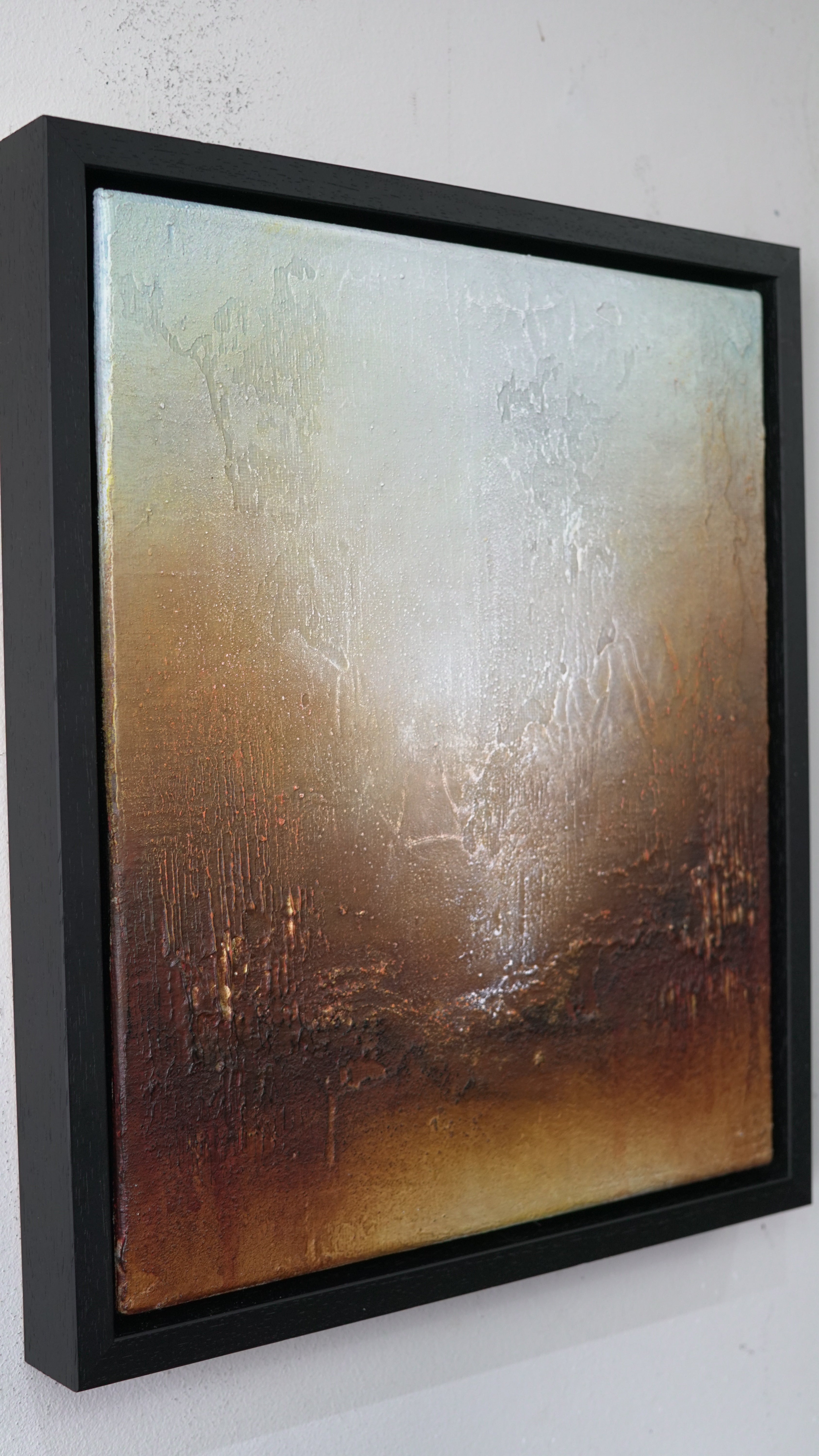 Framed abstract canvas wall art hanging on a white wall, featuring a textured gradient from pale cream and silver at the top to warm brown and bronze tones at the bottom, enclosed in a black frame.