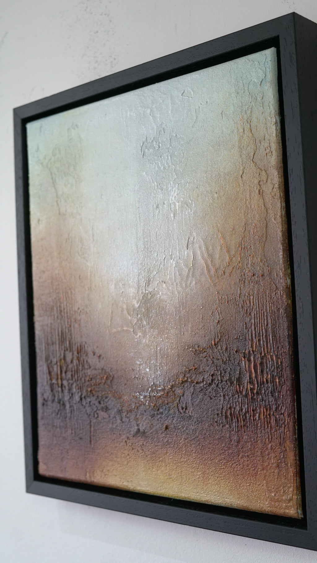 A framed abstract textured painting with a black frame, hanging on a white wall. The artwork features a gradient of cream, beige, brown, and dark brown tones with a metallic sheen.