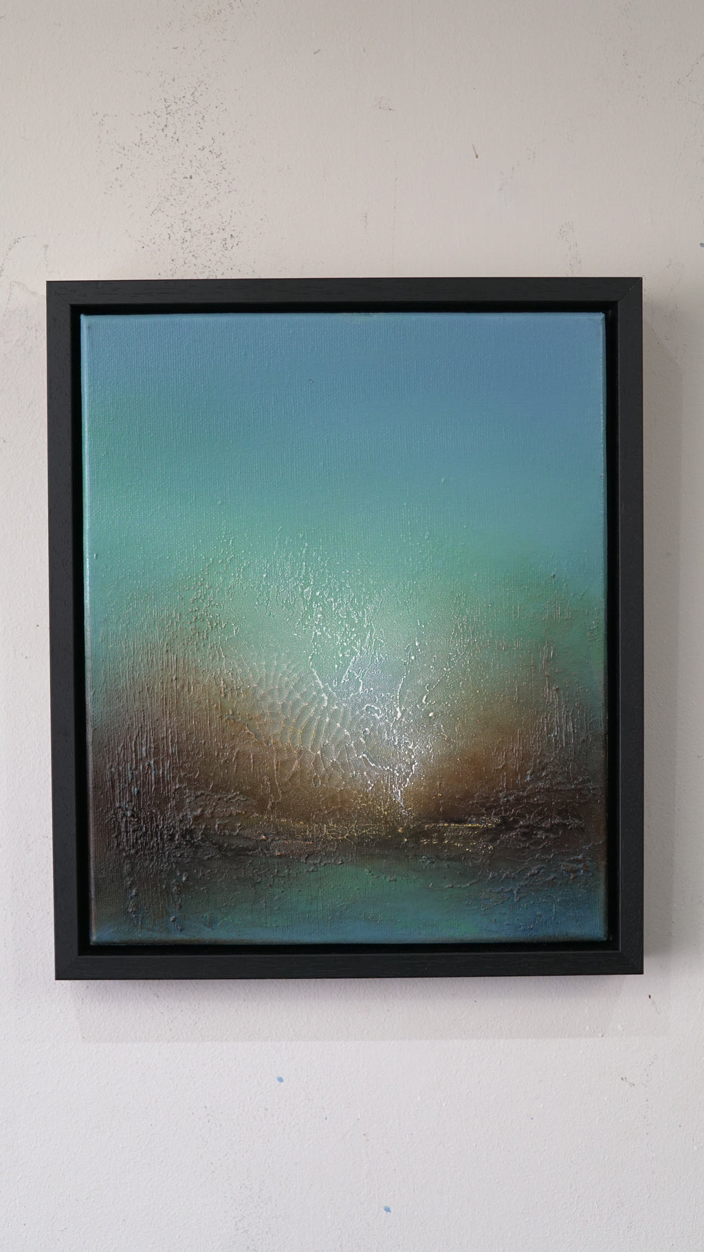 A framed abstract landscape painting featuring a textured gradient of teal, blue, and brown tones, housed in a simple black frame and mounted on a white wall.