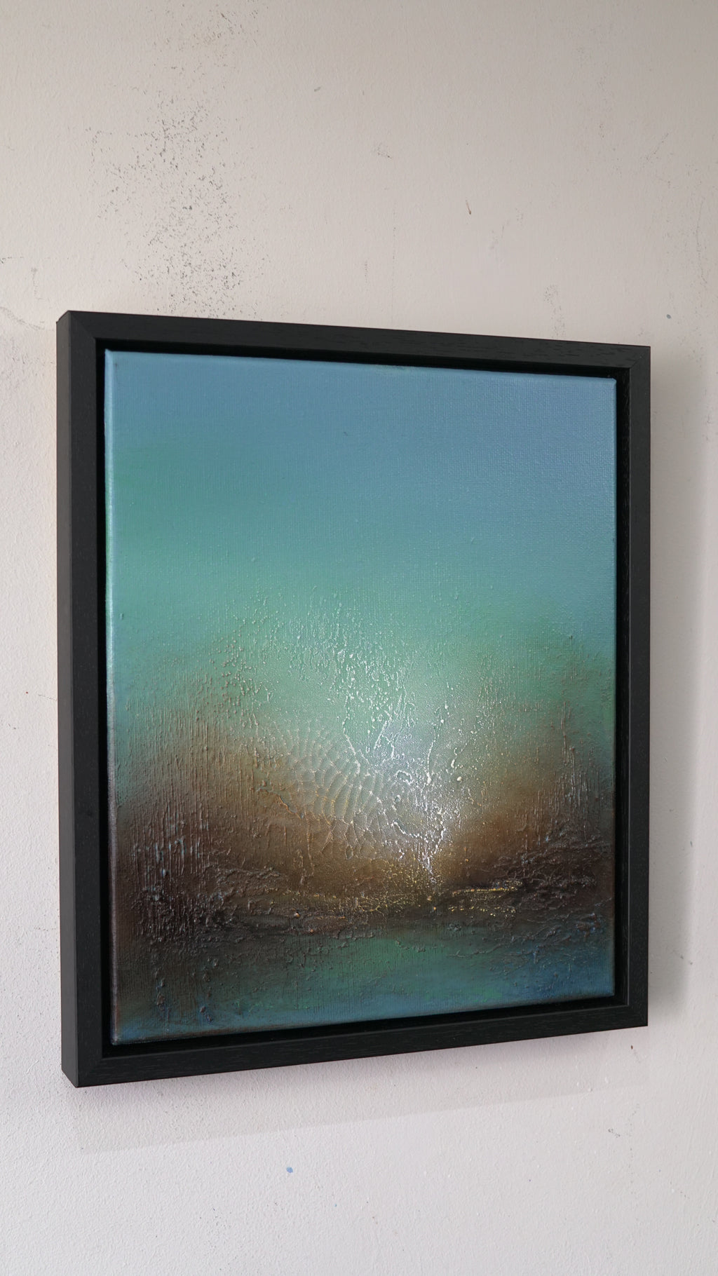 A framed abstract landscape painting hanging on a textured white wall. The artwork features a gradient of teal, green, and brown tones with a textured, wet-looking finish, enclosed in a thick black frame.