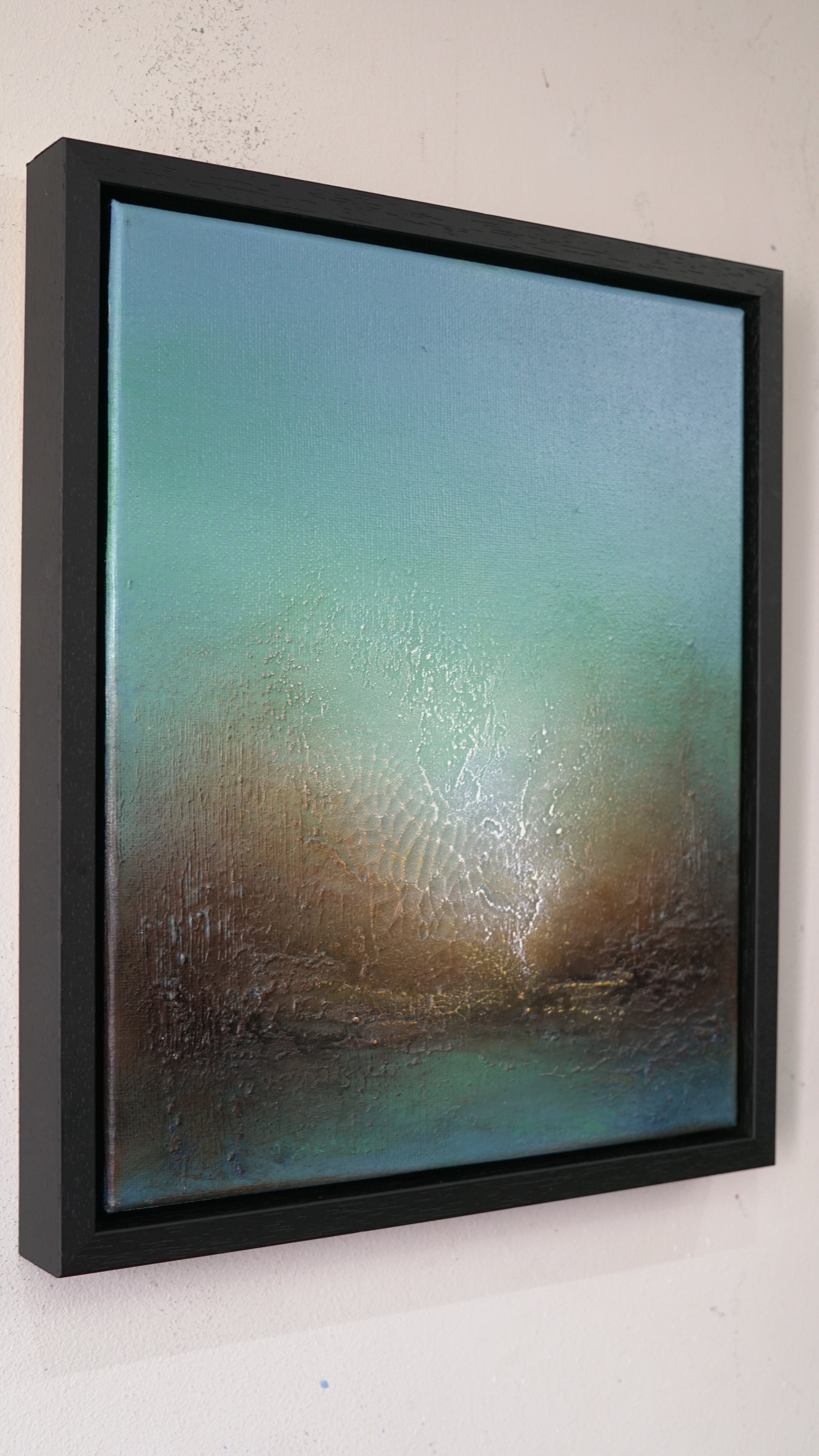 A framed abstract landscape painting featuring a textured teal and brown sky over a dark blue horizon, housed in a thick black frame and mounted on a white wall.