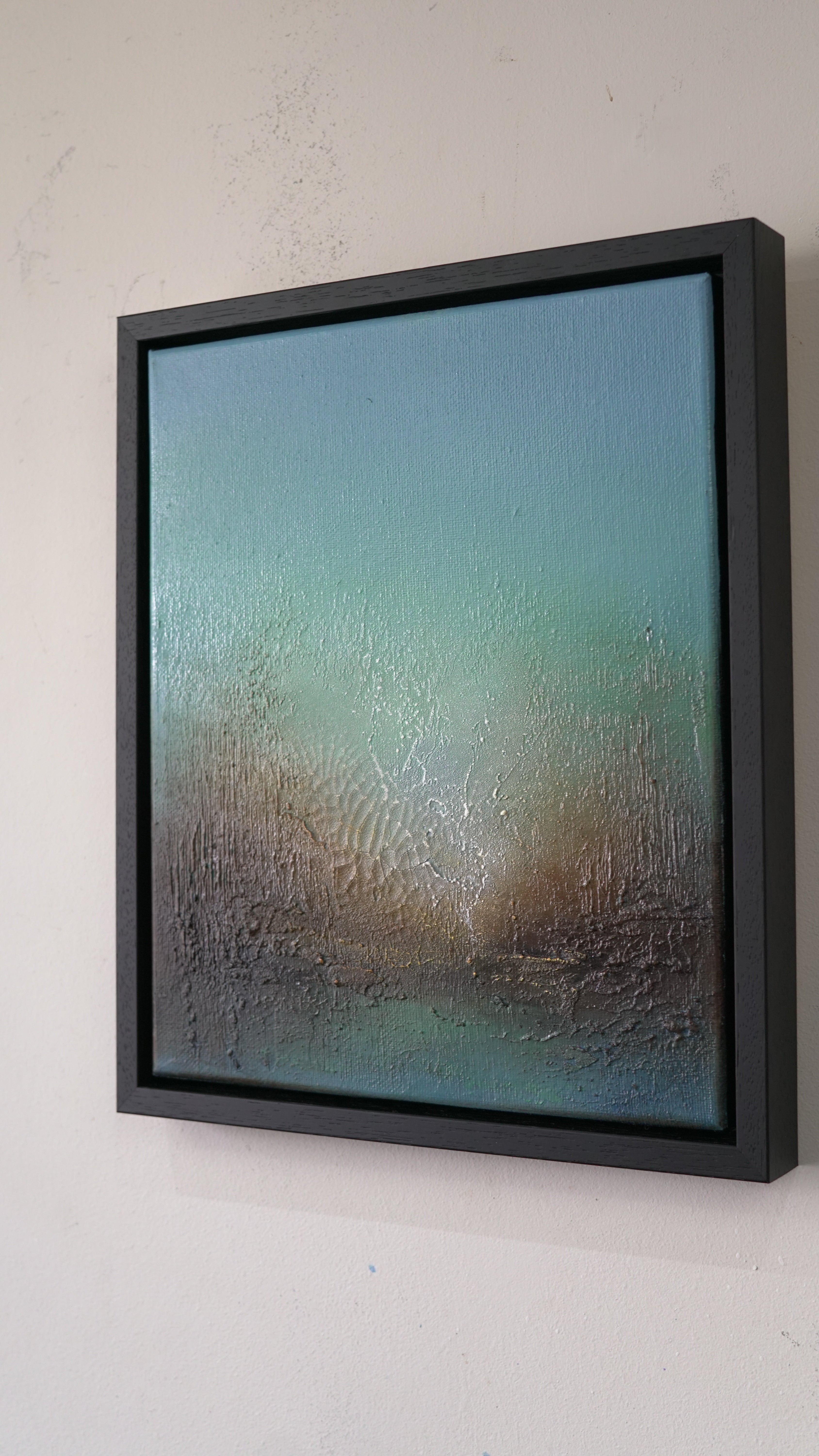 A framed abstract canvas painting with a textured, wet-looking surface, featuring a gradient of teal, blue, and brown tones, housed in a thick black frame and mounted on a light beige wall.