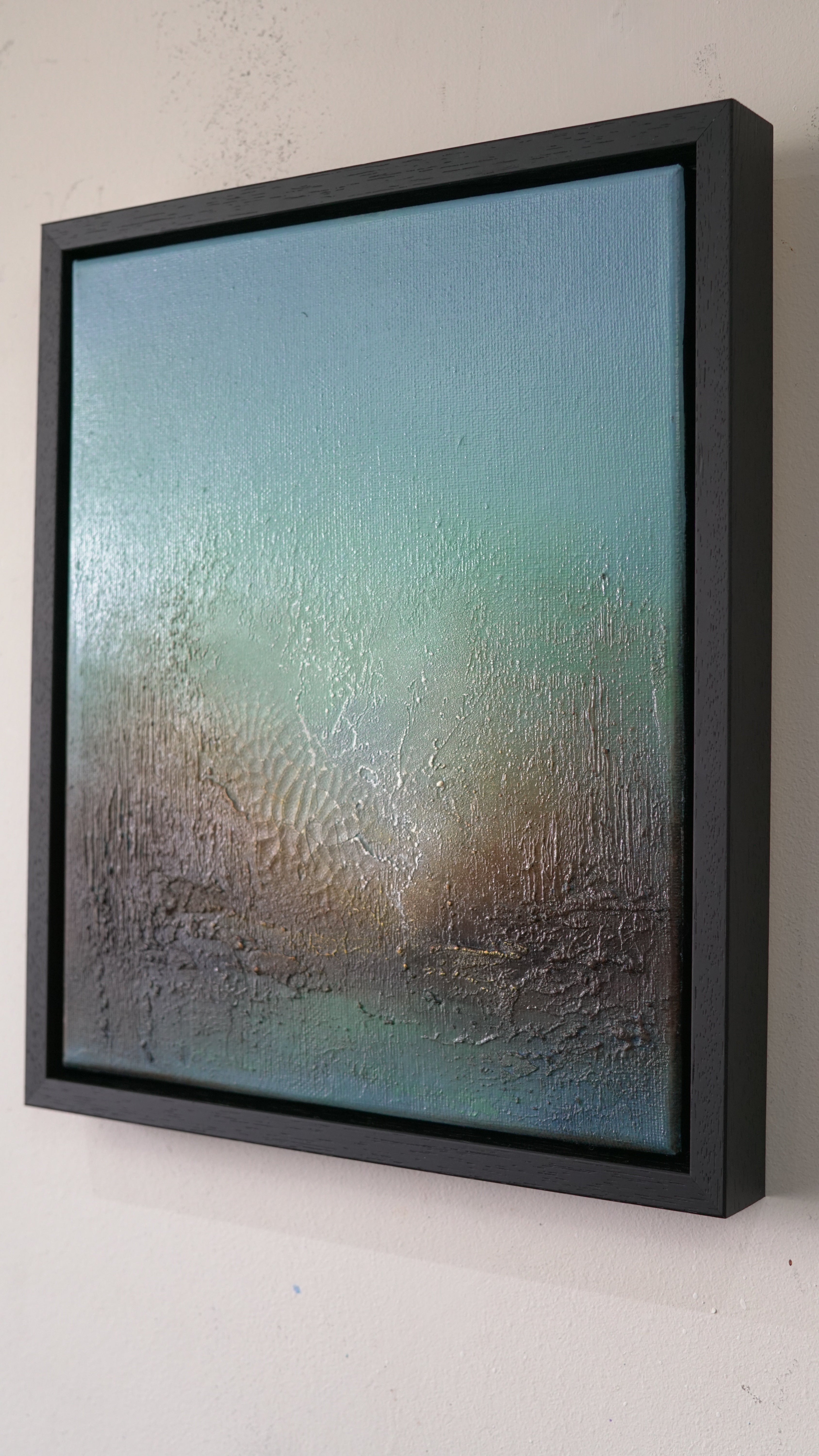A framed abstract canvas painting with a textured, condensation-like surface, featuring a gradient of teal, blue, and brown tones, housed in a thick black frame and mounted on a white wall.