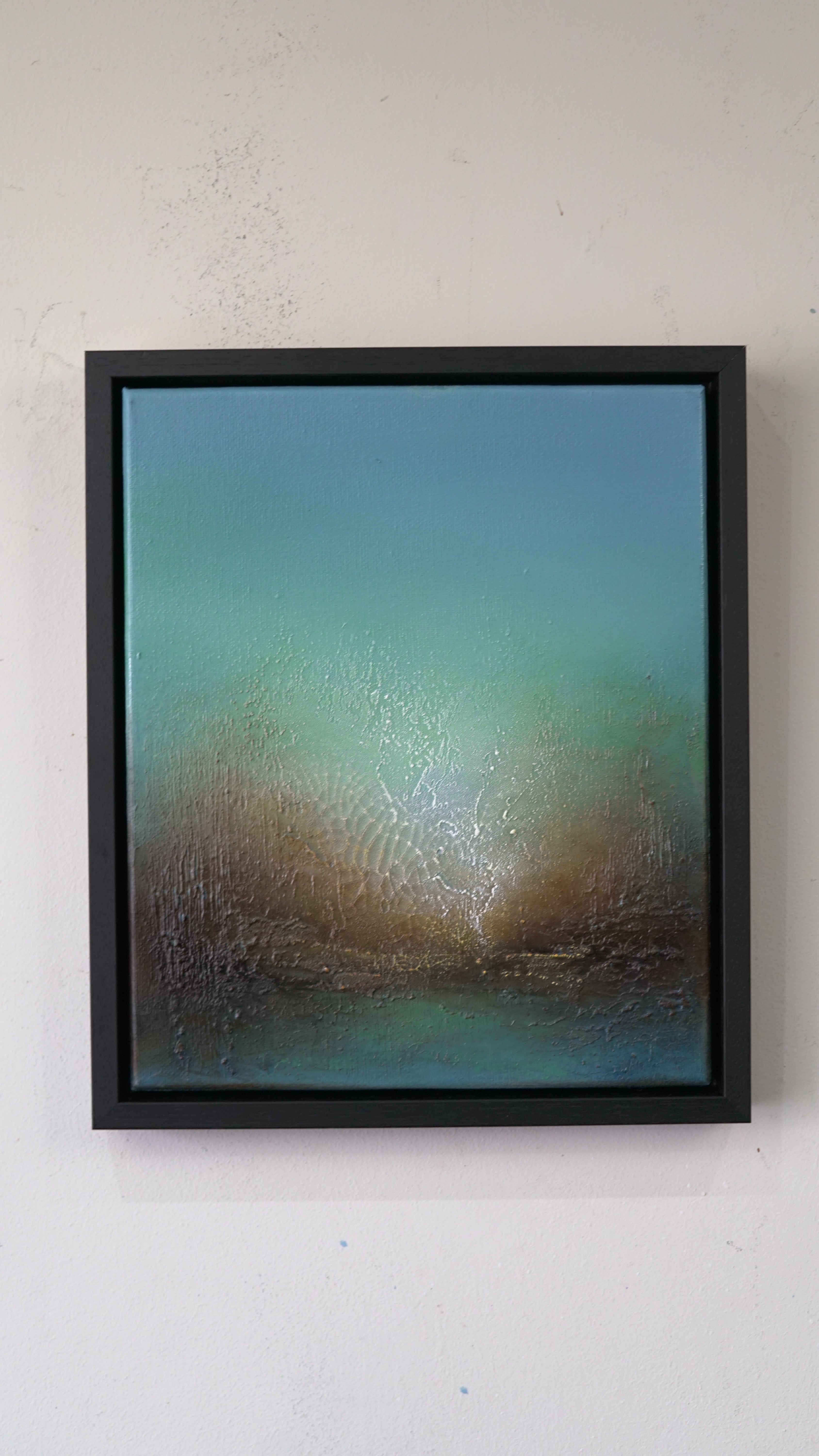 A framed abstract landscape painting featuring a gradient of teal, green, and brown tones with textured details, housed in a black frame and mounted on a light gray wall.