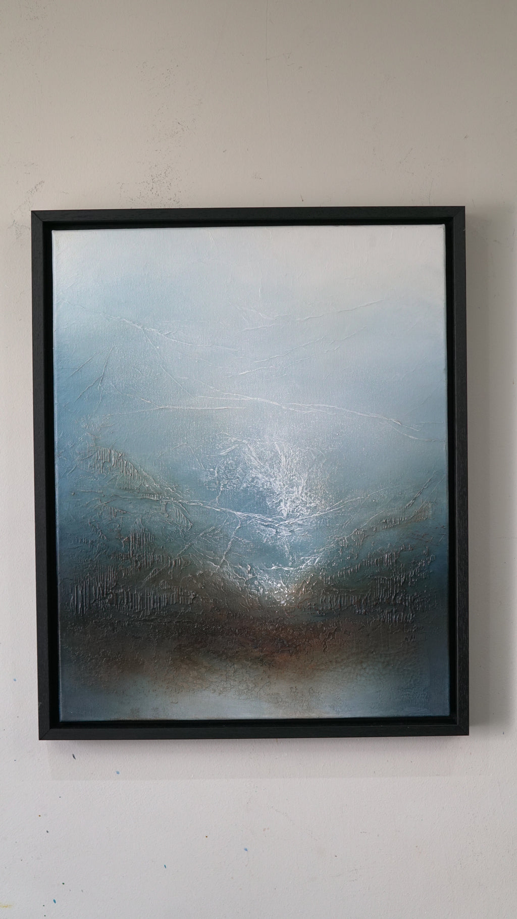 A framed abstract landscape painting featuring textured blue, gray, and brown tones, housed in a simple black frame and hung on a light-colored wall.