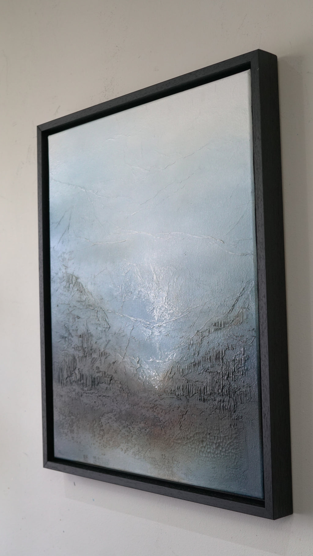 A framed abstract landscape painting featuring textured gray and blue tones, housed in a thick black floating frame and mounted on a light beige wall.