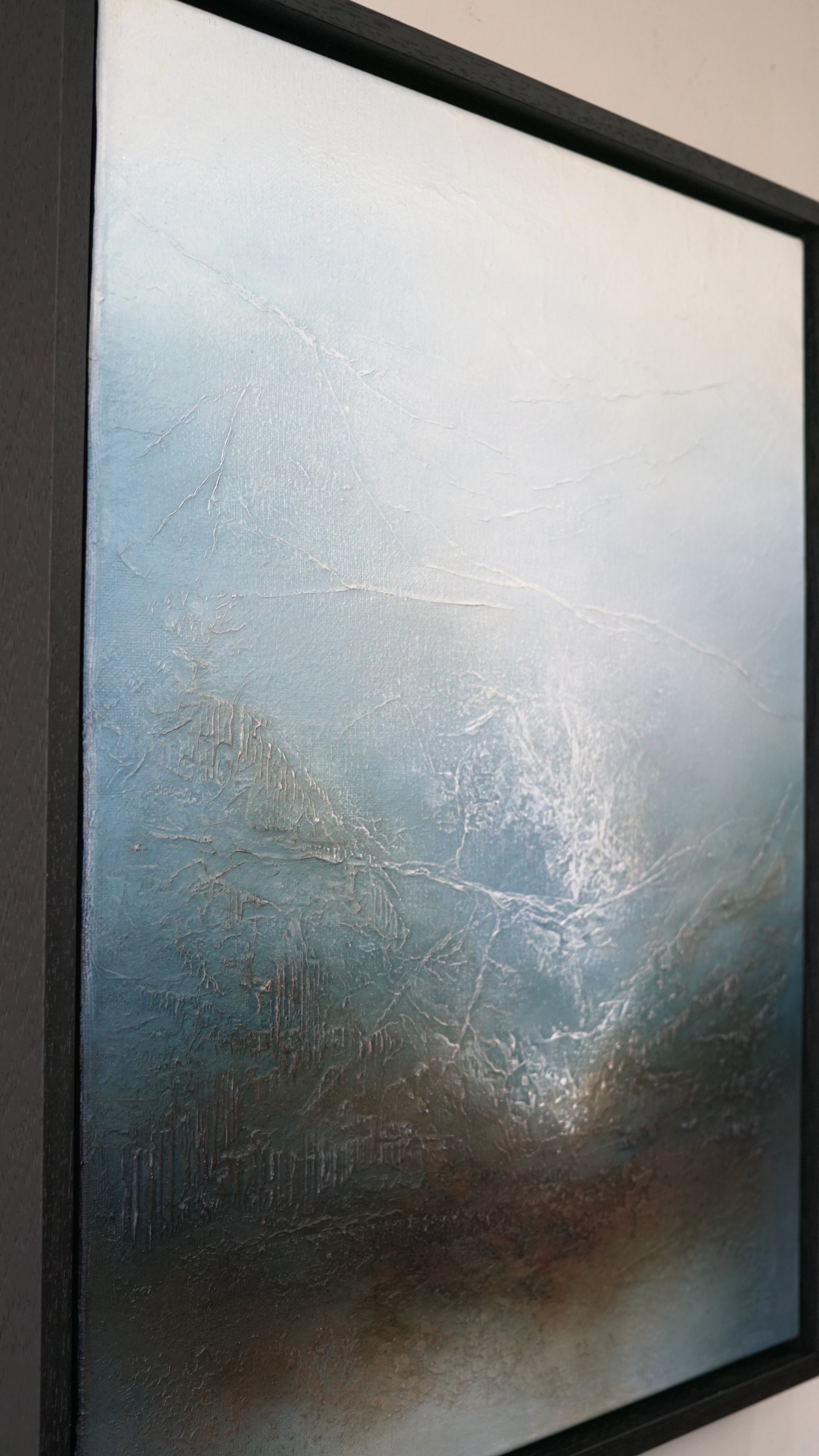 An abstract framed painting featuring textured brushstrokes in shades of blue, gray, and brown, enclosed in a thick black frame.