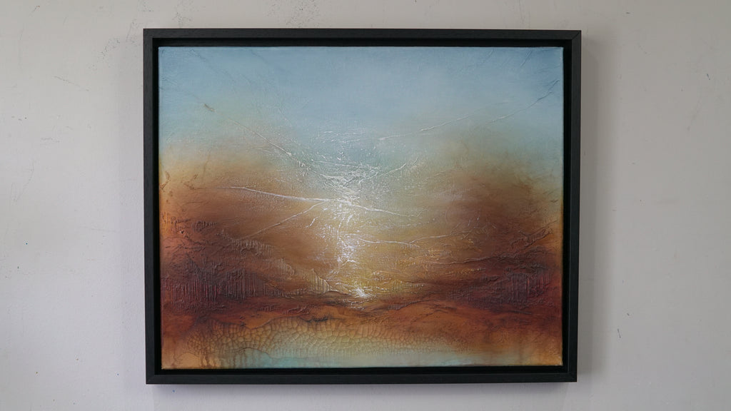 A framed abstract landscape painting hanging on a light gray wall. The artwork features a textured sky gradient from pale blue at the top to warm brown and orange tones near the bottom, with a bright glowing horizon.