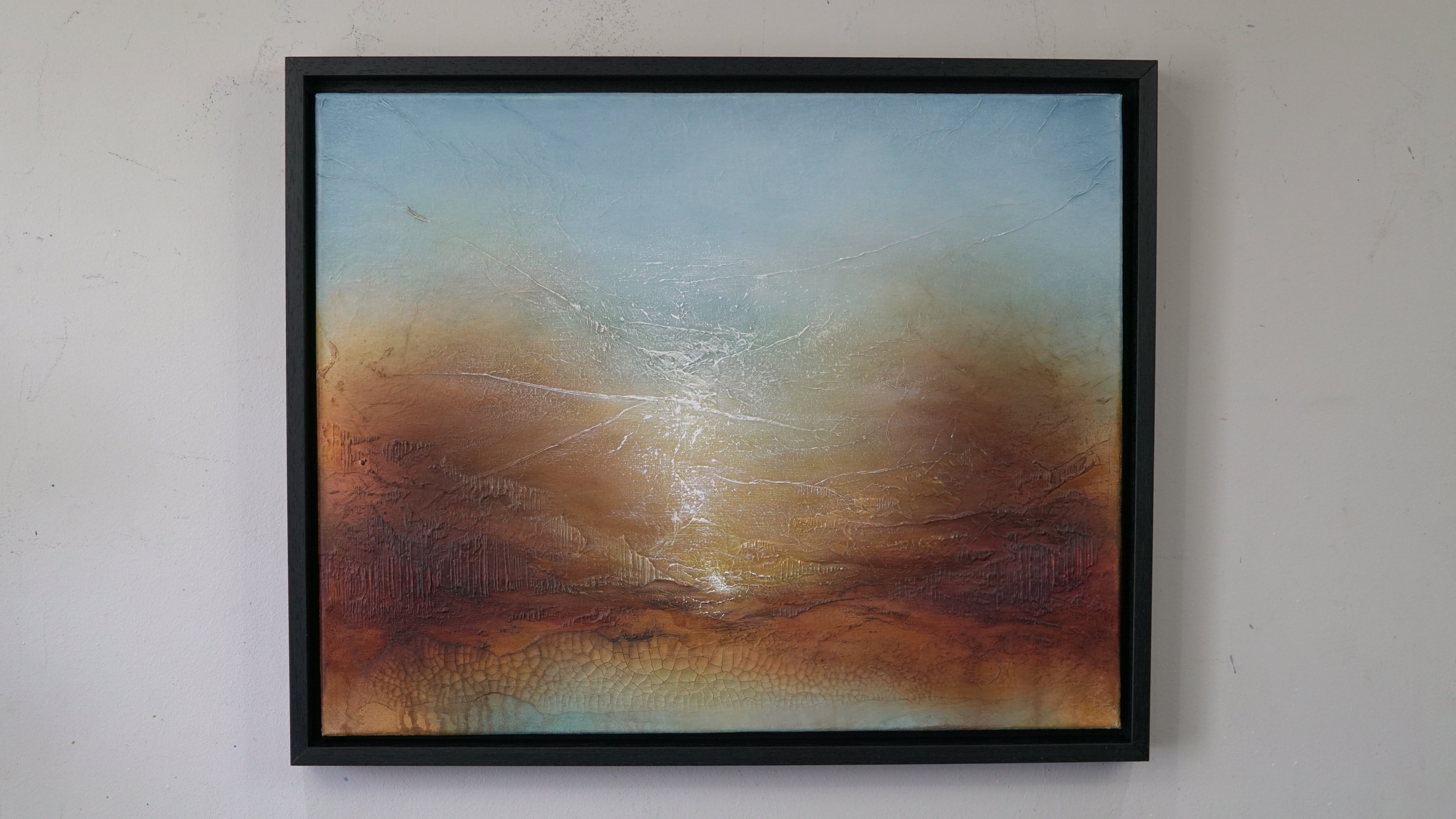 A framed abstract landscape painting hanging on a light gray wall. The artwork features a textured sky gradient from pale blue at the top to warm brown and orange tones near the bottom, with a bright glowing horizon.