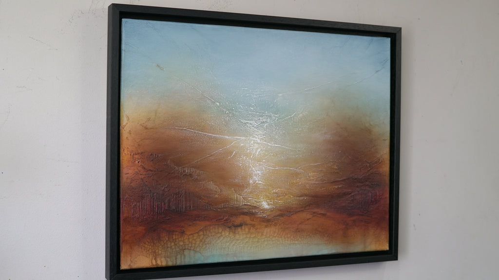 An abstract framed painting hanging on a white wall. The artwork features a textured blend of blue, orange, brown, and gold tones with a bright central glow, enclosed in a black frame.