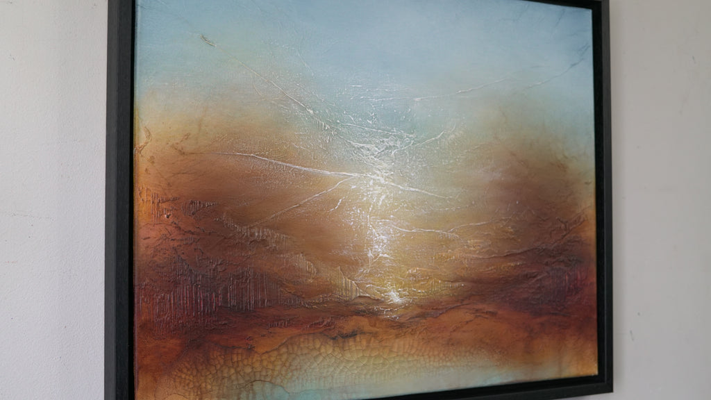 A framed abstract landscape painting hanging on a white wall. The artwork features a textured sky in shades of blue and orange, with a bright sun-like glow in the center and a dark brown earthy foreground.