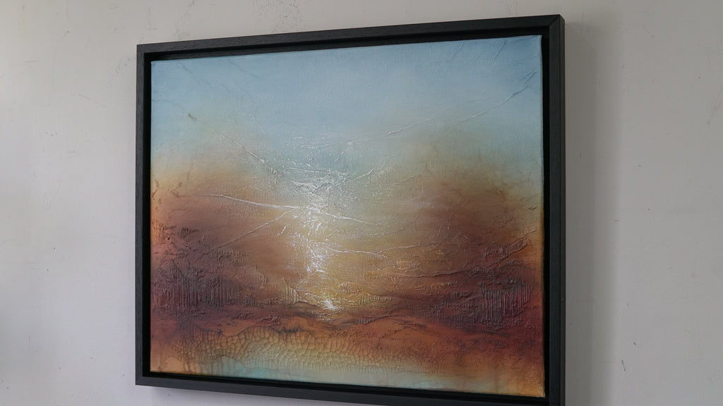 A framed abstract landscape painting hanging on a white wall. The artwork features a textured sky gradient from light blue at the top to warm brown and orange tones near the bottom, with a bright glowing center.
