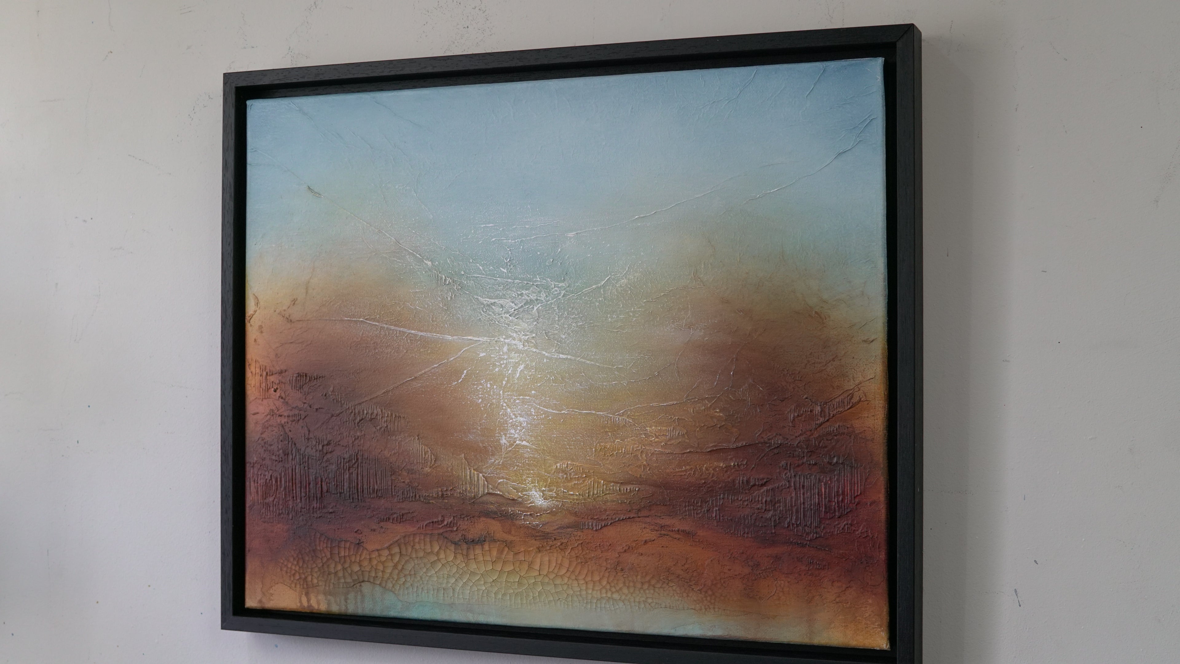 A framed abstract landscape painting hanging on a white wall. The artwork features a textured sky gradient from light blue at the top to warm brown and orange tones near the bottom, with a bright glowing center.