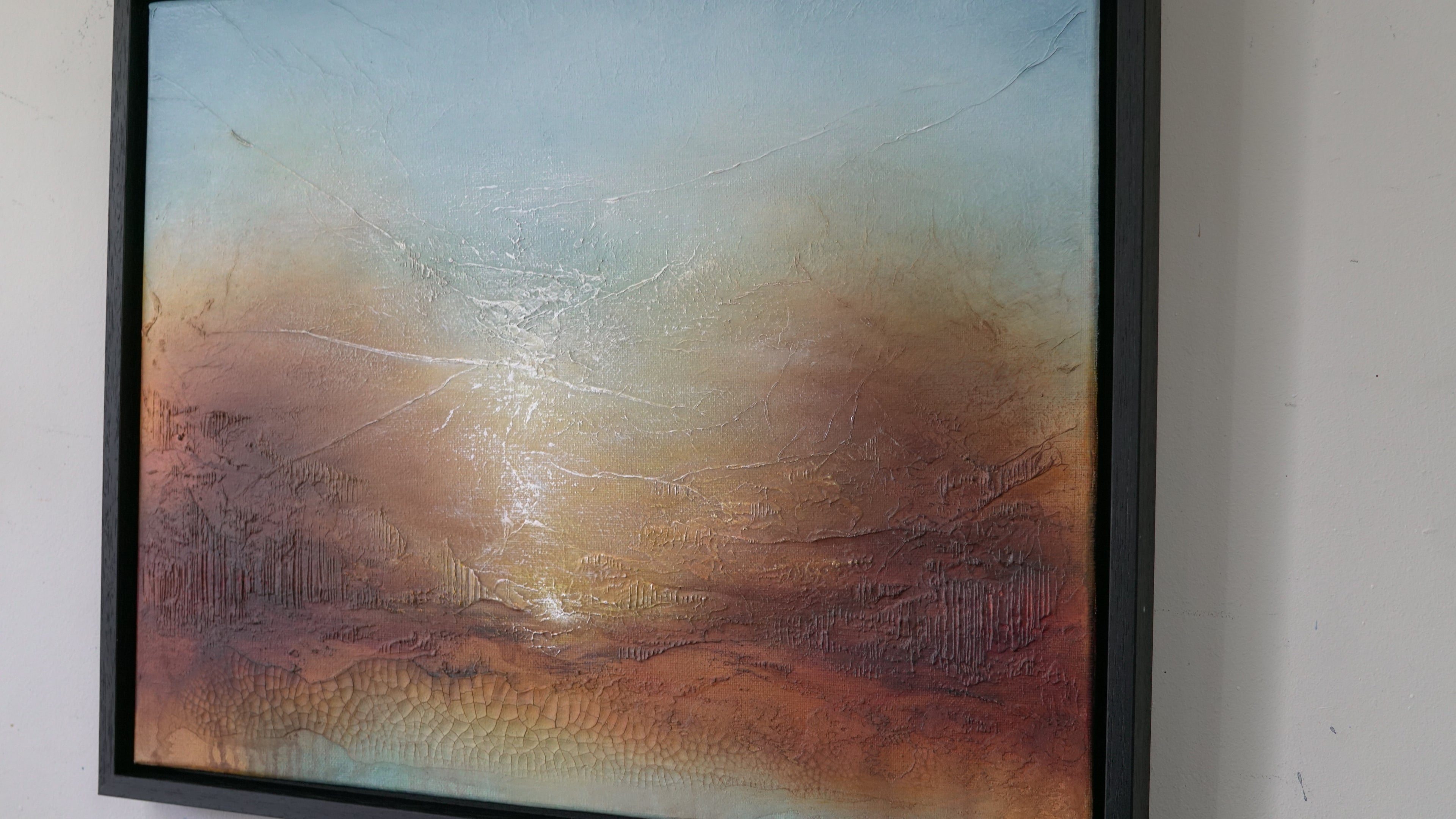 A framed abstract landscape painting featuring textured brushstrokes in shades of brown, orange, beige, and light blue, housed in a black frame and displayed on a white wall.