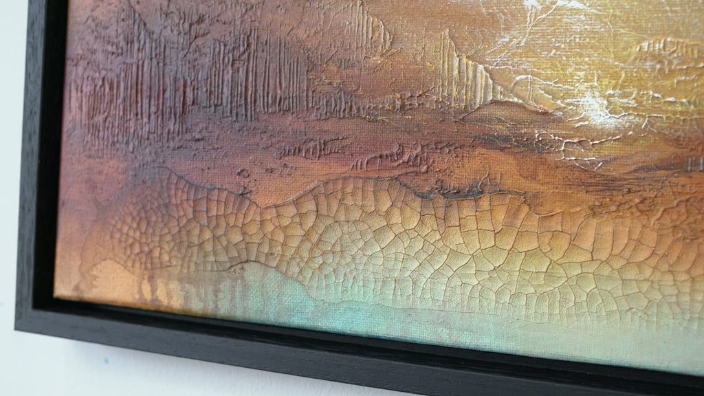 Close-up of a framed abstract textured painting with a black frame, showing warm brown, orange, and gold tones with a crackled surface and a hint of pale blue-green at the bottom.