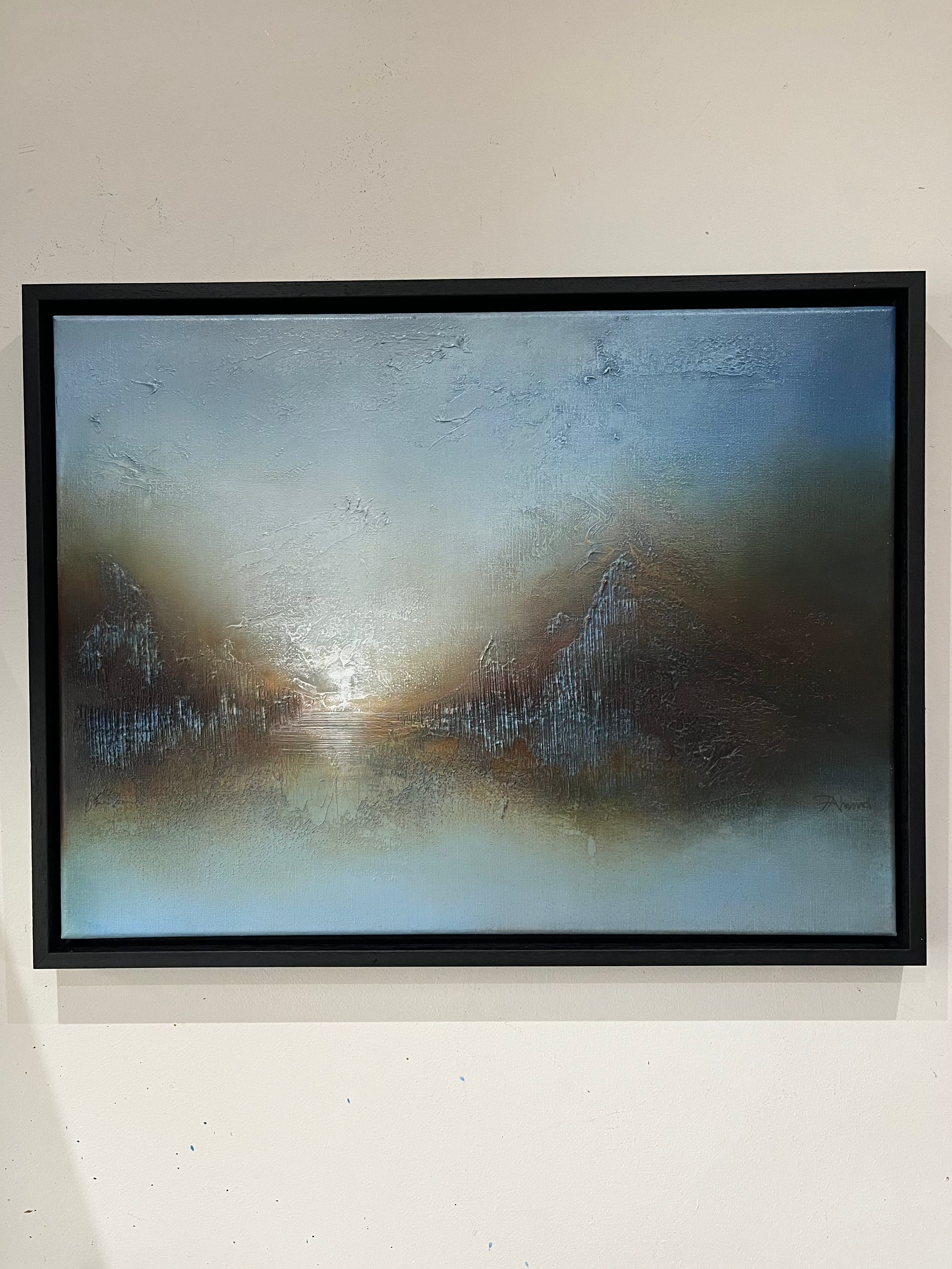 A framed abstract painting featuring a textured, smoky composition with shades of blue, gray, brown, and white, housed in a simple black frame and mounted on a light beige wall.