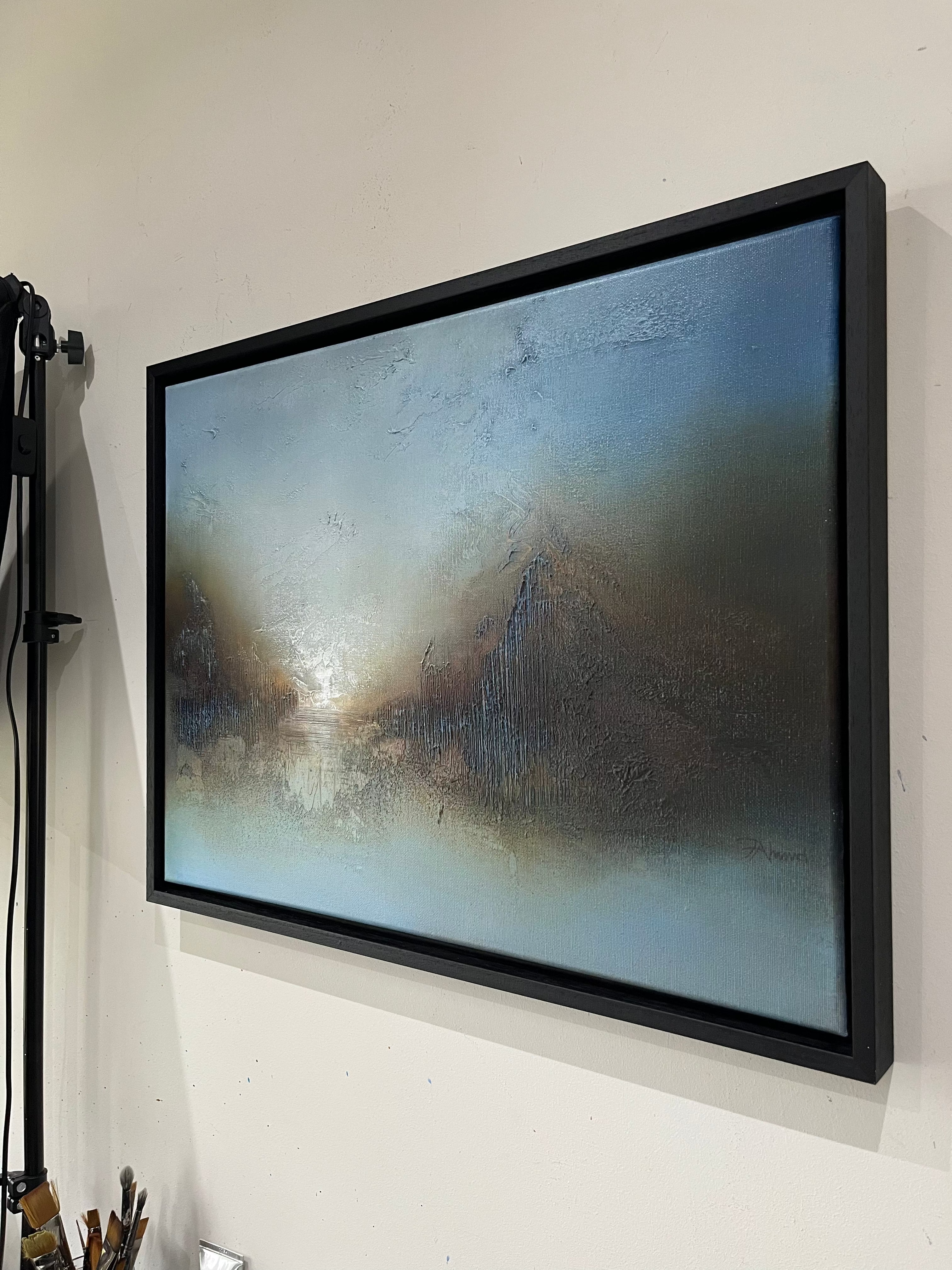 A framed abstract painting with a textured, metallic-looking surface in shades of blue, gray, and brown, housed in a thick black frame and hanging on a white wall.