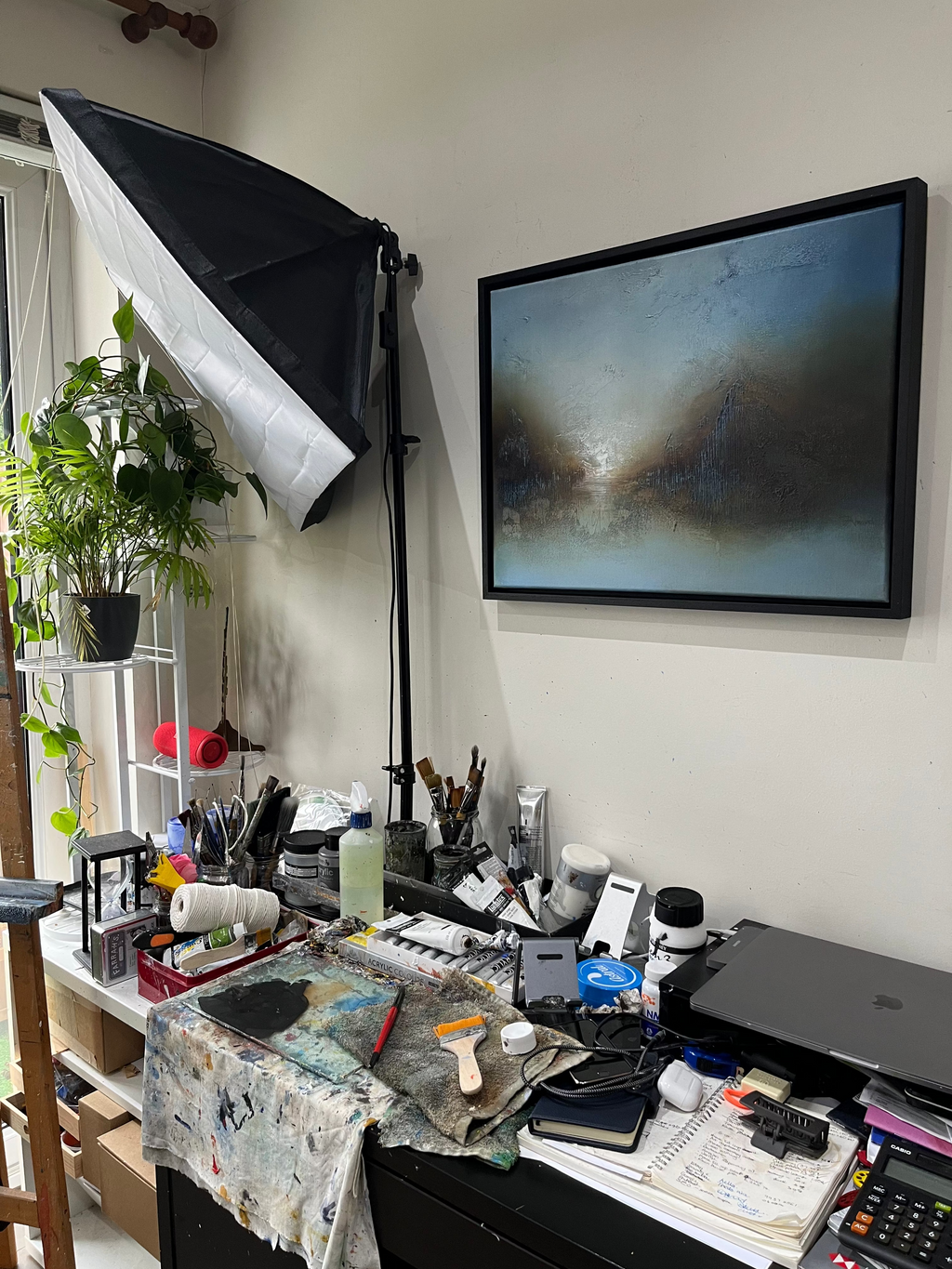 A framed abstract landscape painting hangs on a white wall in a studio or art workspace. The artwork features blue, gray, and brown tones.
