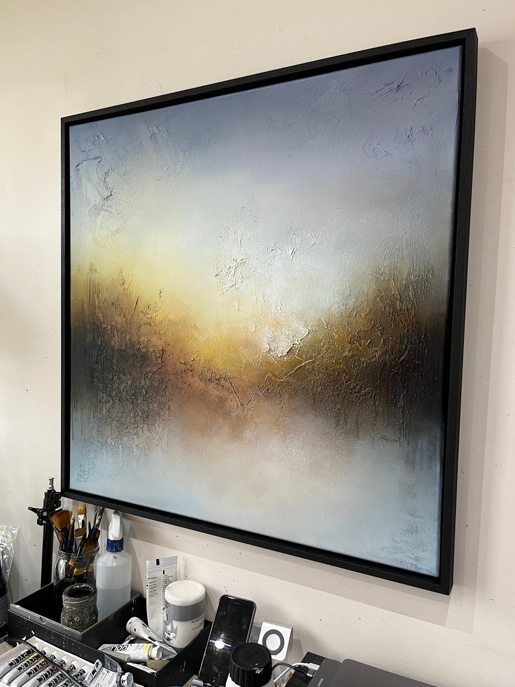 My Light framed abstract painting hanging above a desk with art supplies, in blue, grey, gold and brown tones