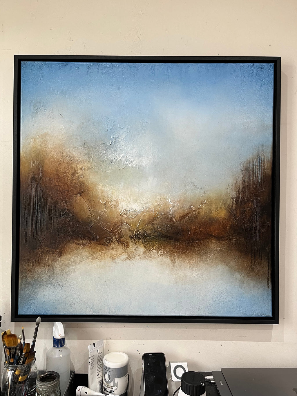 A framed abstract landscape painting hanging on a beige wall above a desk with office supplies. The painting features a textured, impressionistic design with a blue sky, warm brown and gold hills, and a white foreground, enclosed in a black frame.