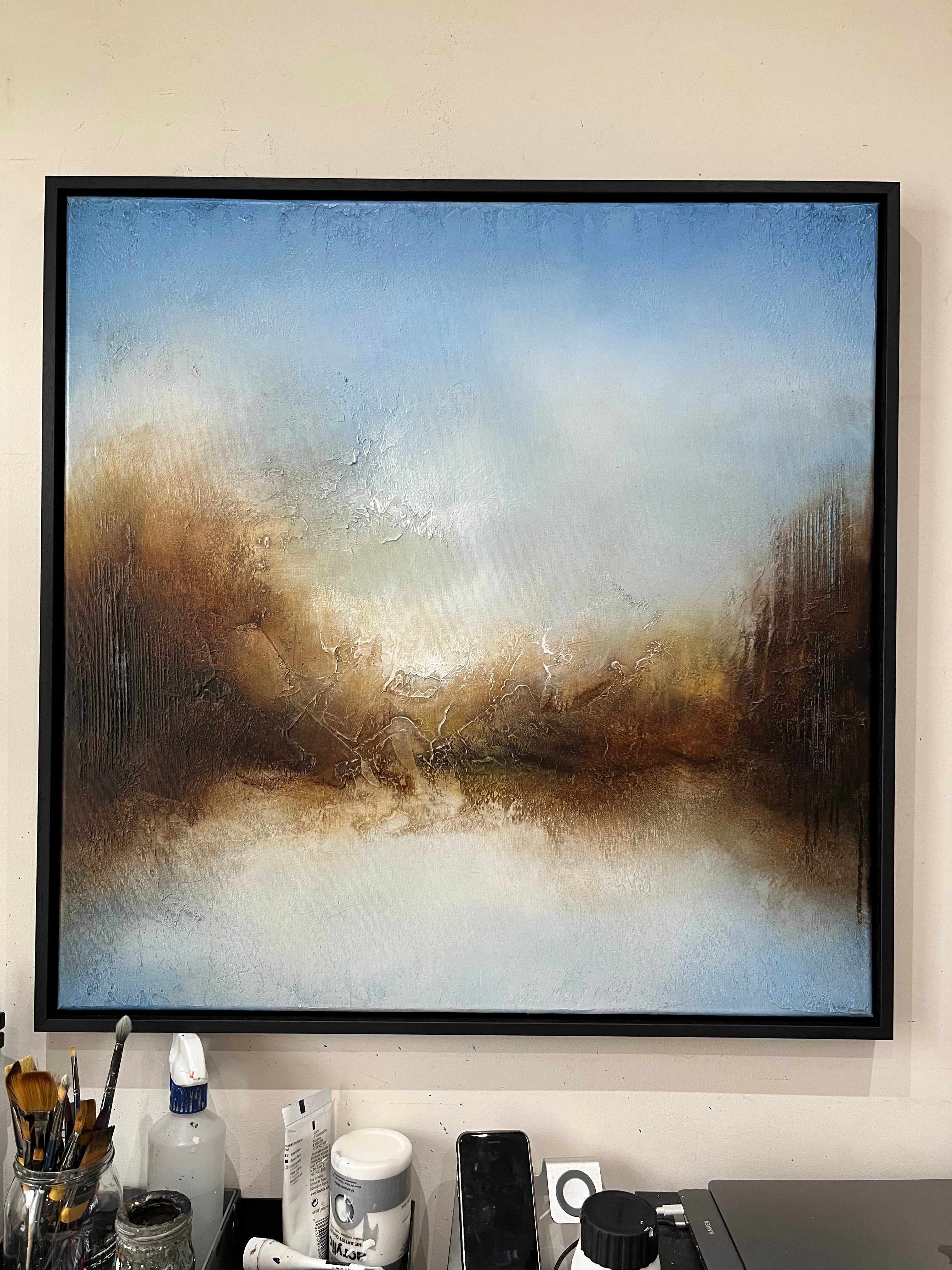 A framed abstract landscape painting hanging on a beige wall above a desk with office supplies. The painting features a textured, impressionistic design with a blue sky, warm brown and gold hills, and a white foreground, enclosed in a black frame.