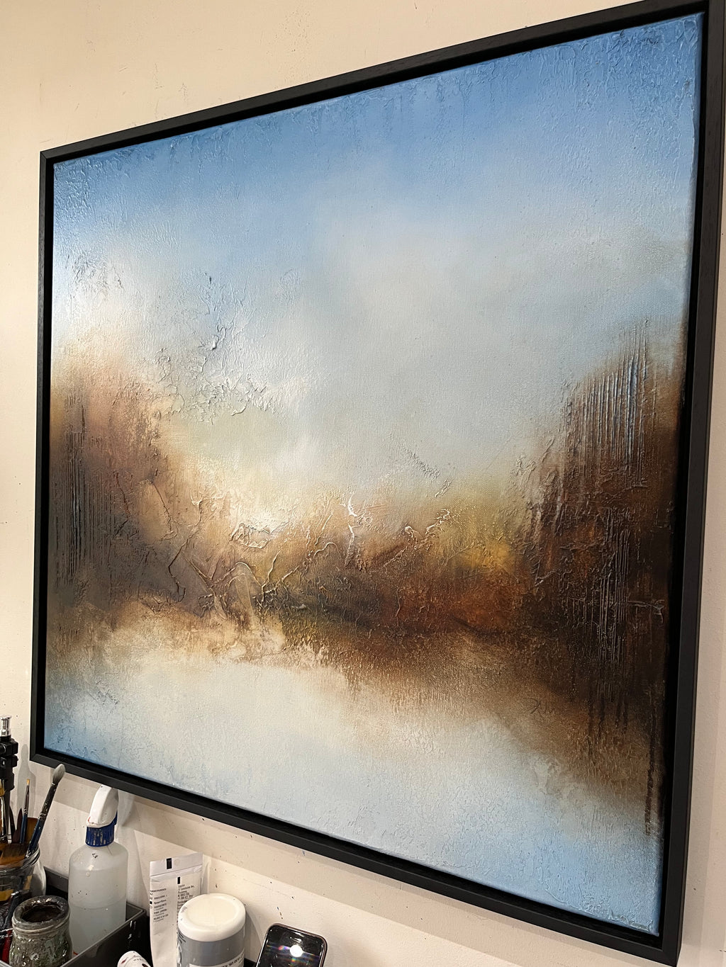 A large framed abstract landscape painting with textured brushstrokes in blue, beige, brown, and white, hanging on a wall above a countertop with bottles and brushes.