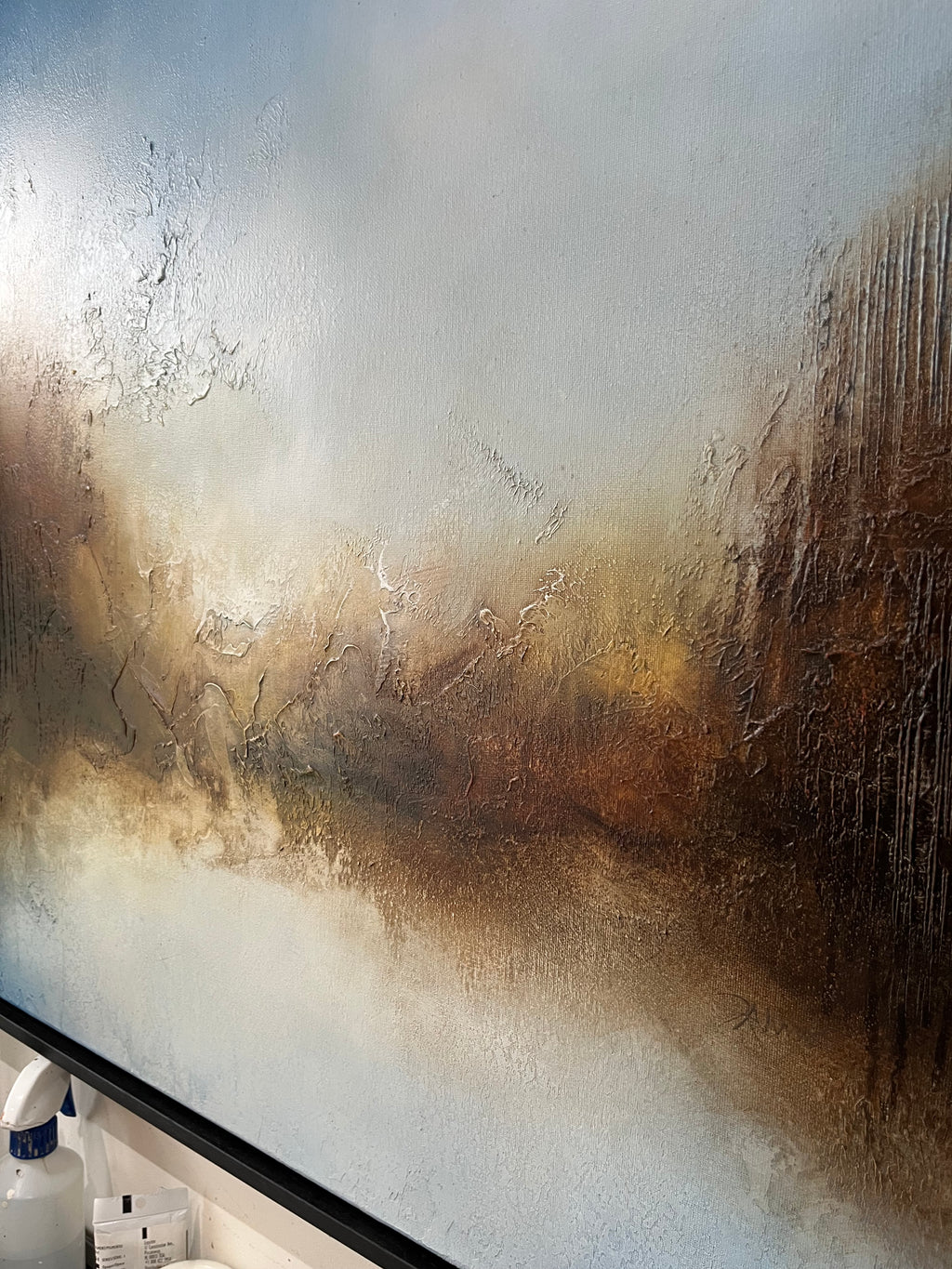Abstract textured wall art featuring a landscape-like scene with layered brushstrokes in blue, white, brown, and gold tones, shown hanging above a dark-framed mirror or glass panel.
