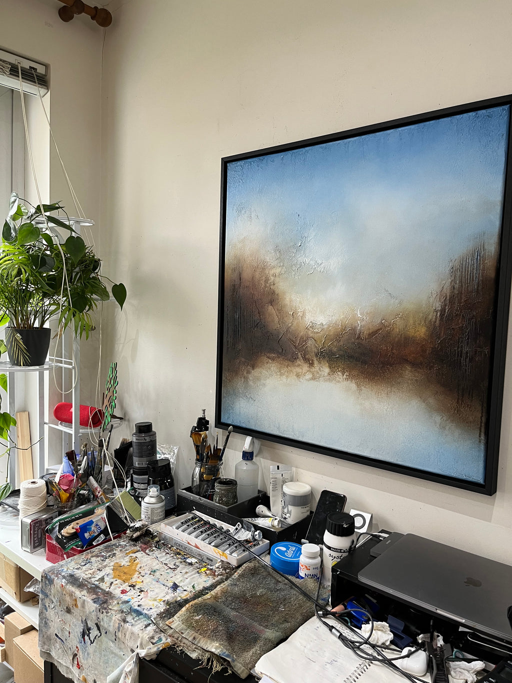A large framed abstract landscape painting with blue, white, and brown tones hangs on a beige wall above a cluttered artist's studio desk with paint tubes, brushes, and art supplies.
