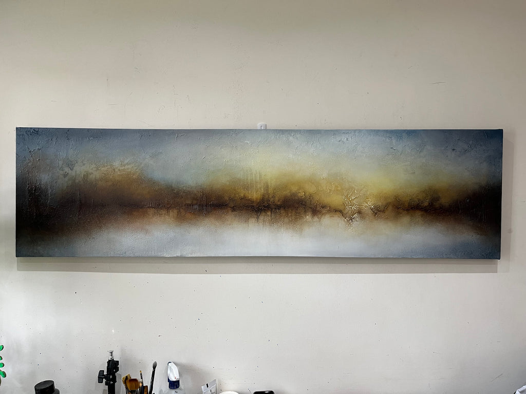 A wide rectangular abstract canvas painting hanging on a light beige wall. The artwork features blended horizontal bands of blue, gray, brown, and warm gold tones, with a textured finish.