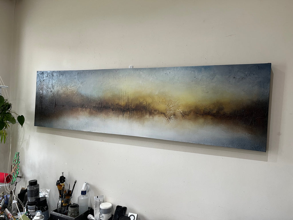 A panoramic abstract canvas painting with textured brushstrokes in blue, gray, gold, and brown, displayed on a plain off-white wall above a desk with art supplies.