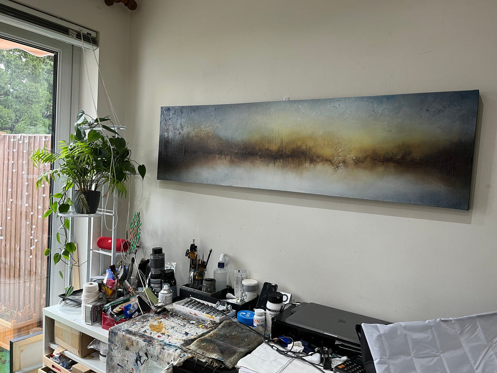 A panoramic abstract canvas painting with a textured landscape of blue, gray, and golden-brown tones, displayed on a wall in a creative studio workspace.