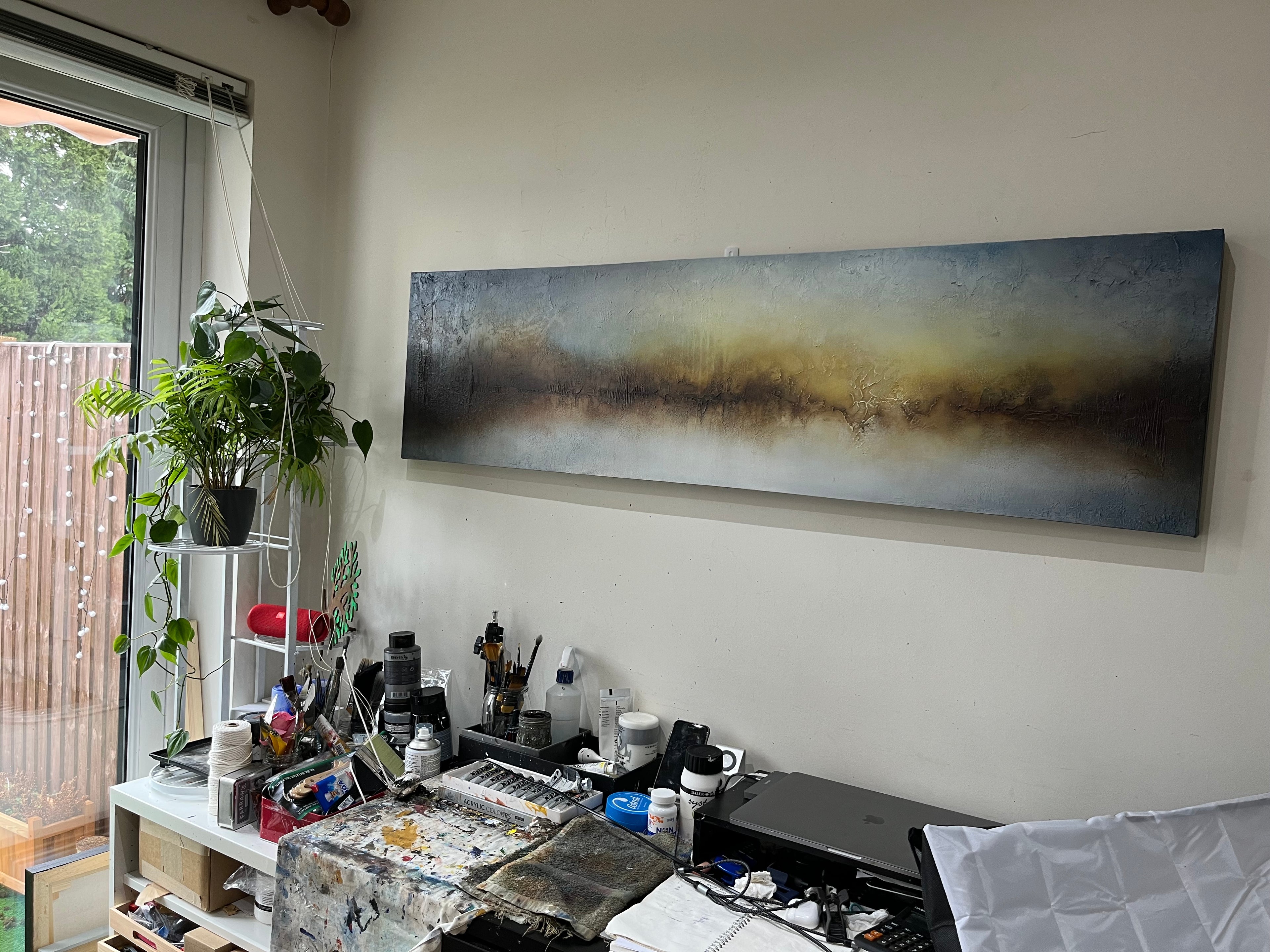 A panoramic abstract canvas painting with a textured landscape of blue, gray, and golden-brown tones, displayed on a wall in a creative studio workspace.