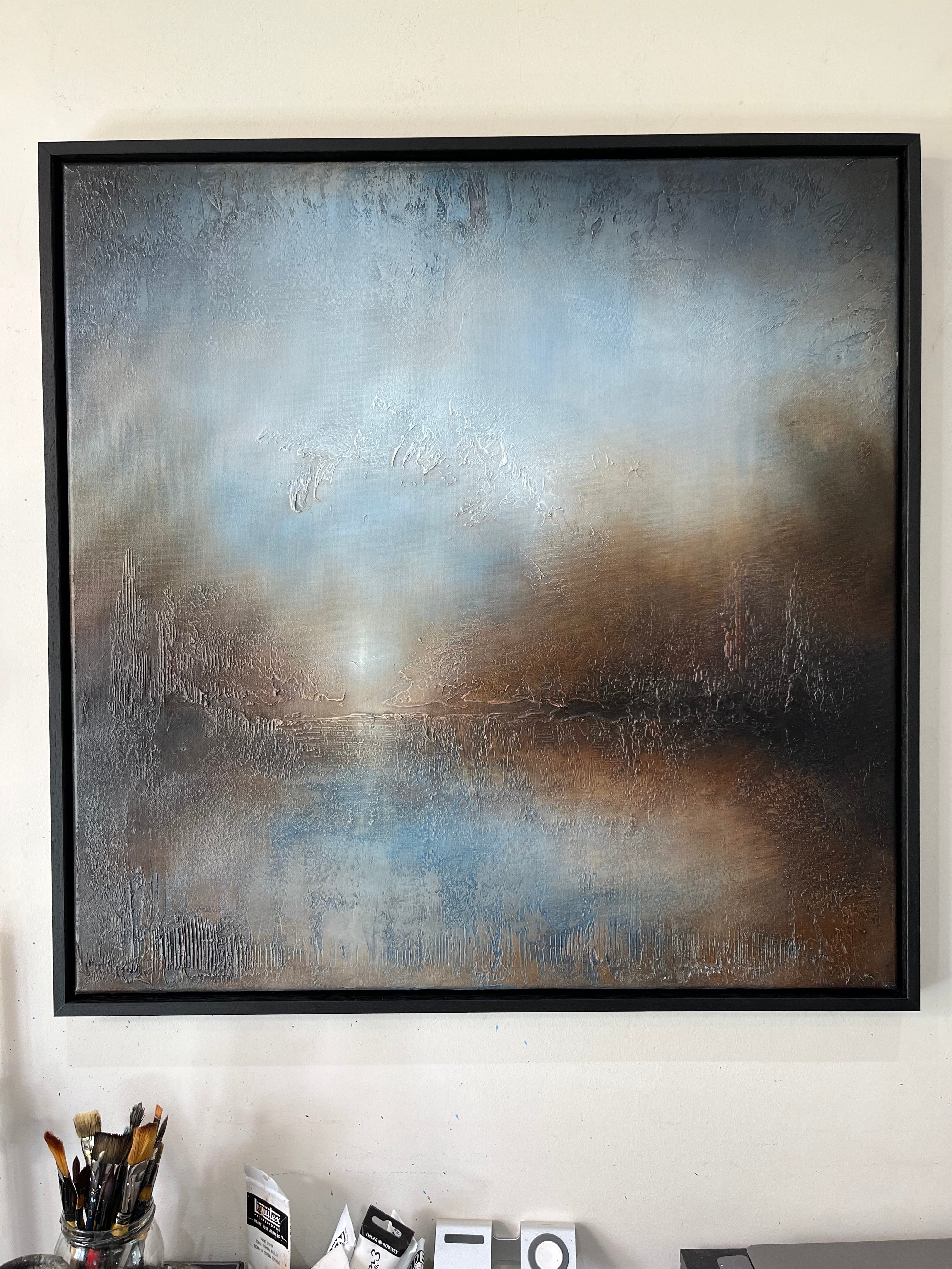 A large framed abstract landscape painting with textured brushstrokes in shades of blue, gray, brown, and white, hanging on a beige wall above a desk with art supplies.