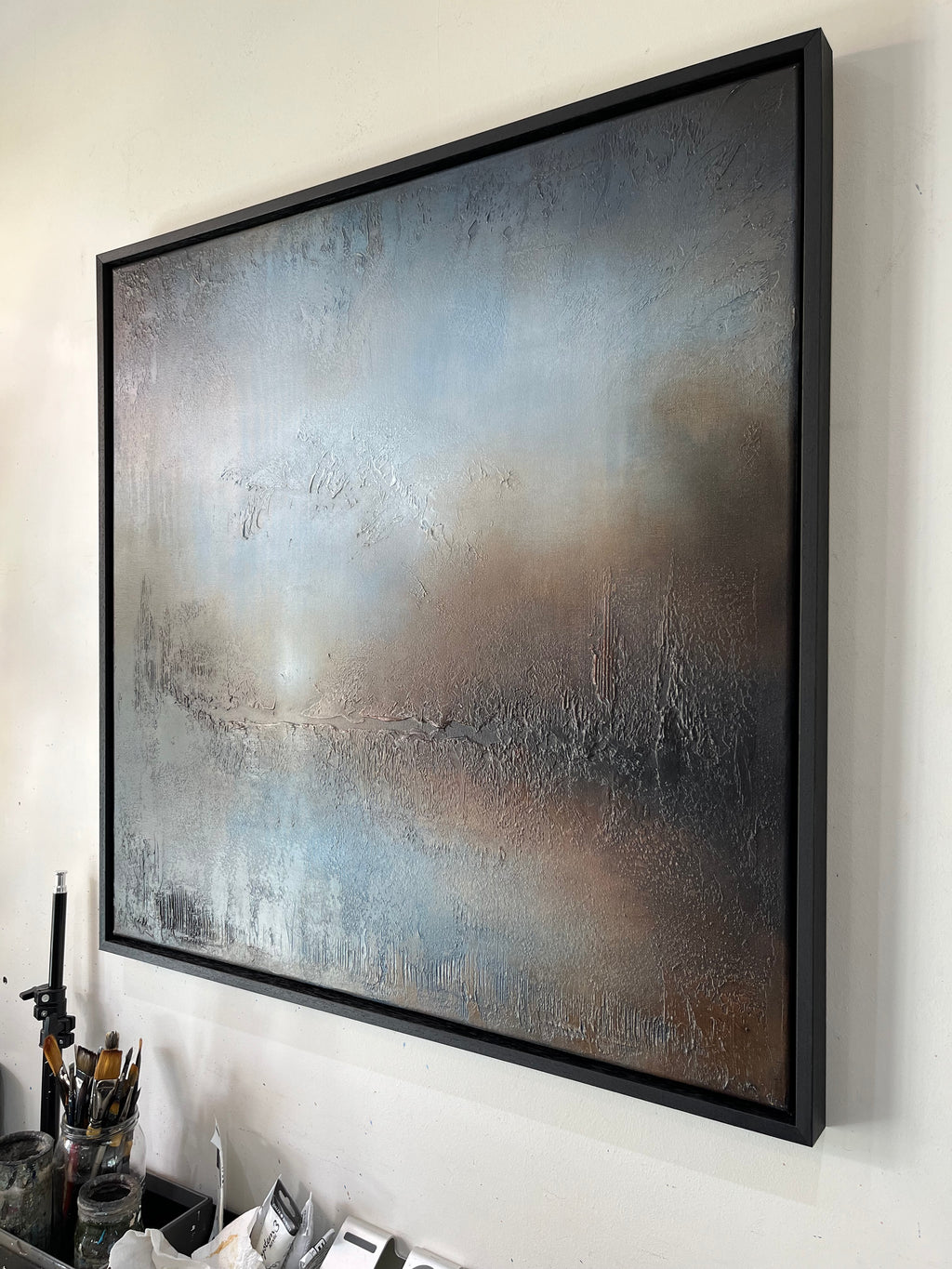 A large framed abstract canvas painting with textured brushstrokes in muted blue, gray, brown, and beige tones, hanging on a white wall above a desk with art supplies.