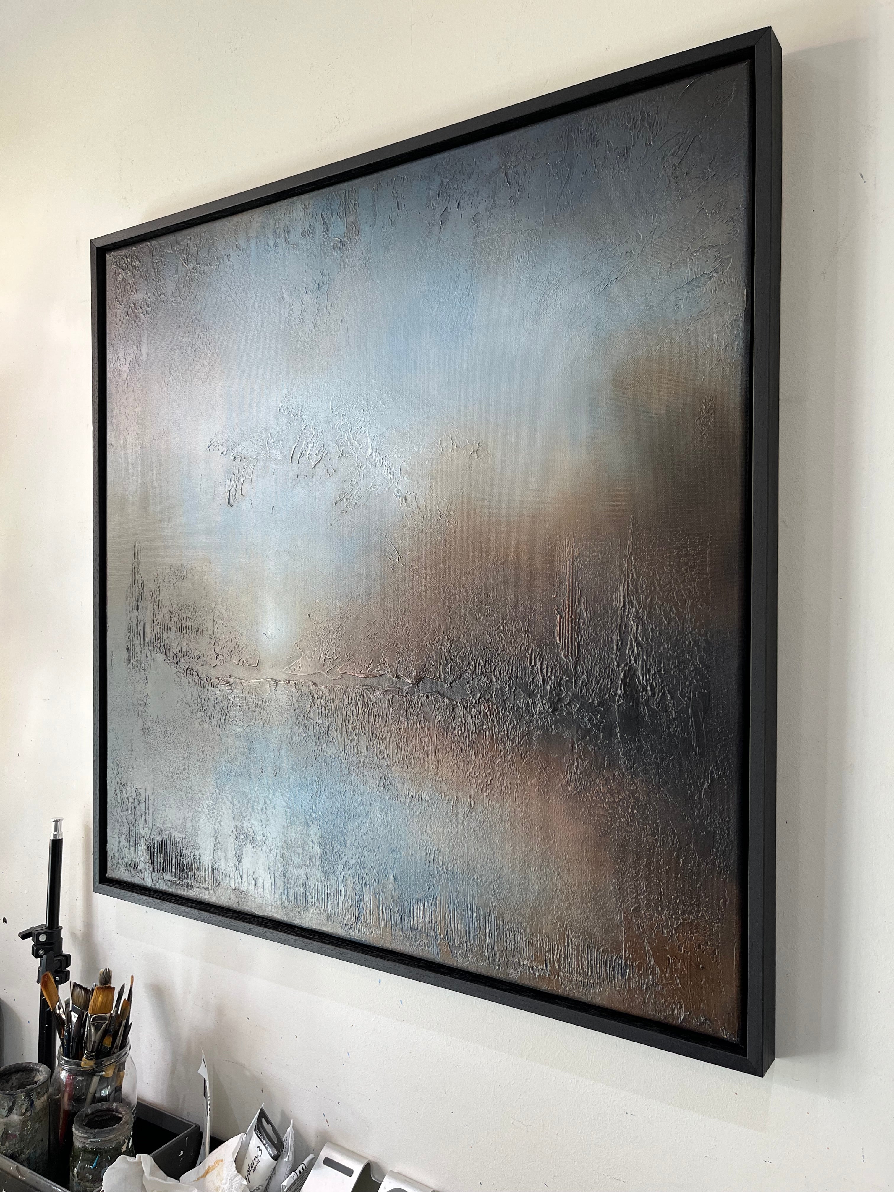 A large framed abstract canvas painting with textured brushstrokes in muted blue, gray, brown, and beige tones, hanging on a white wall above a desk with art supplies.