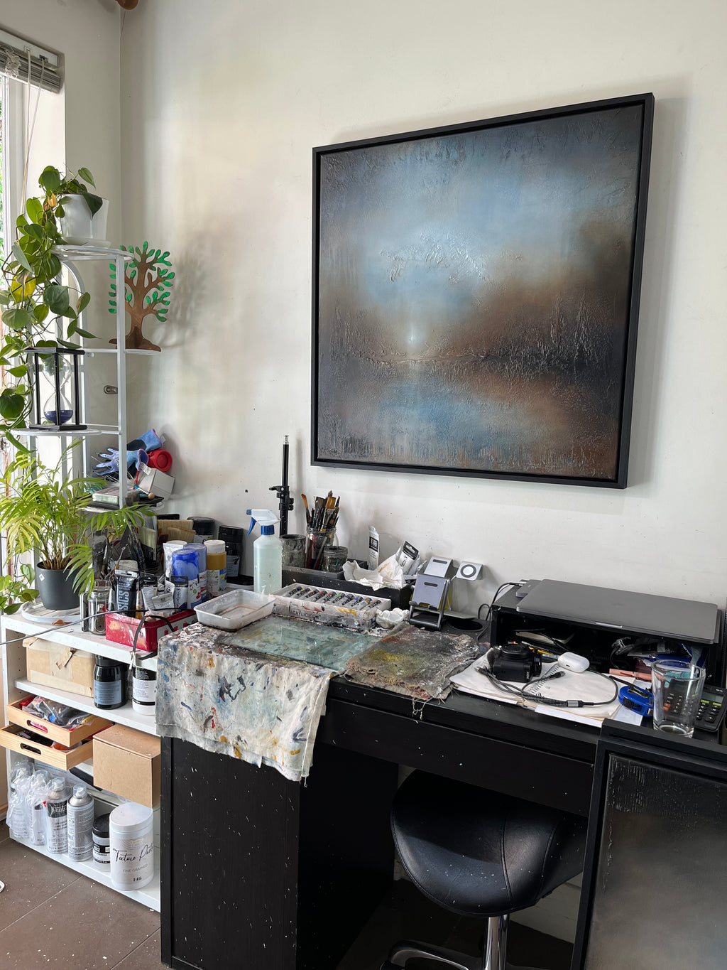 A large framed abstract painting with blue, gray, and brown tones hangs on a white wall above a cluttered artist's studio desk with paint cans, brushes, and art supplies.
